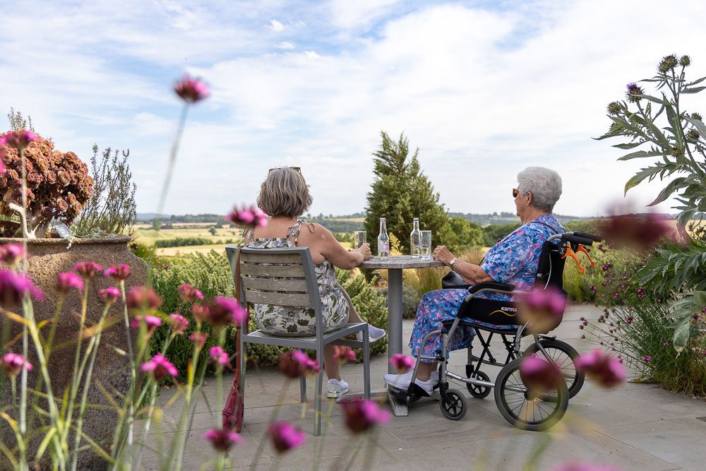 Two people enjoying a garden view from a patio with pink flowers in the foreground and countryside in the distance.