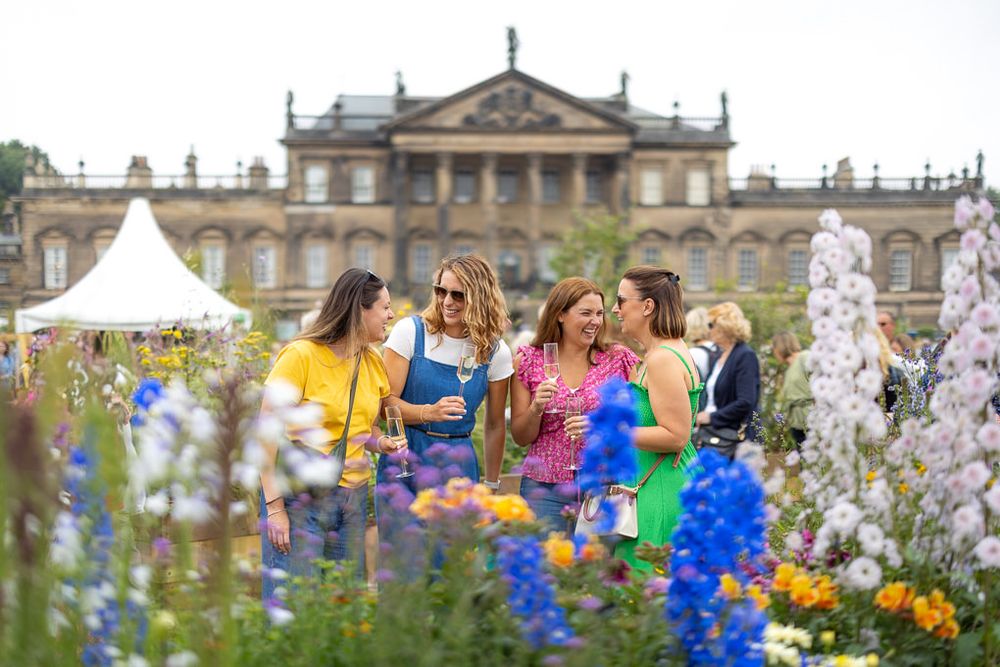 Group enjoying summer drinks and conversation among colorful flower gardens at a historic mansion estate.