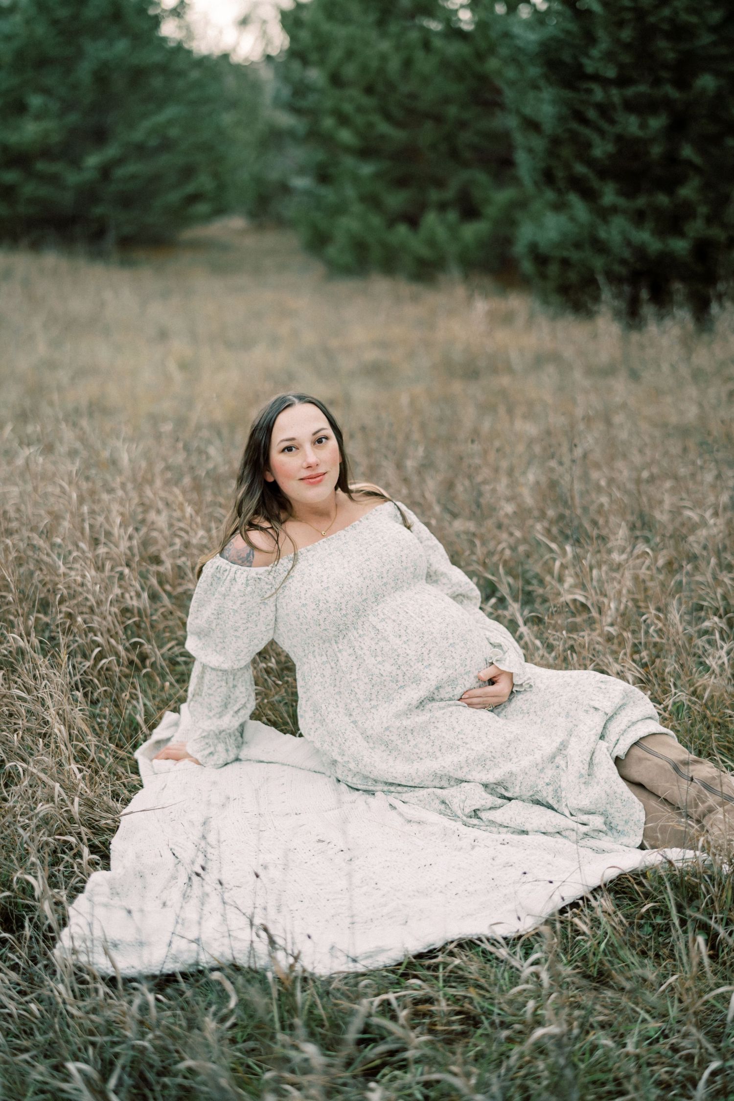 Expectant mother in white lace dress poses gracefully in a field of tall grass at sunset.