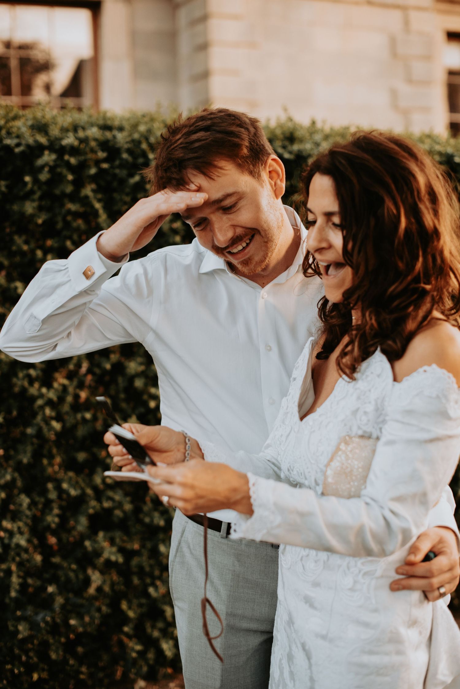 A couple in white wedding attire share a joyful candid moment while looking at a phone outdoors.