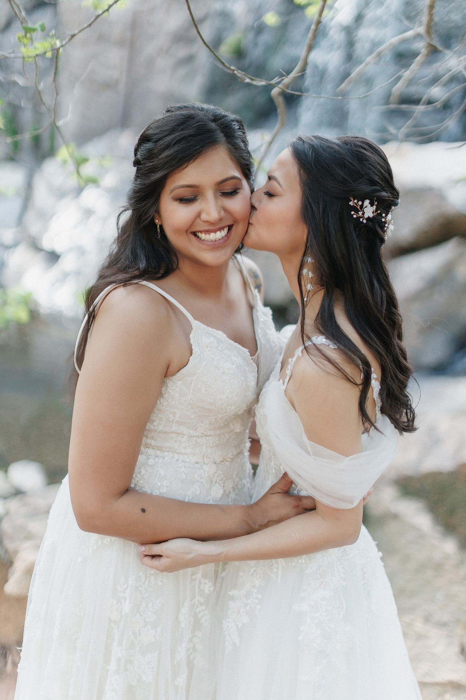 Two brides in white lace wedding dresses share an intimate moment outdoors in a natural setting.