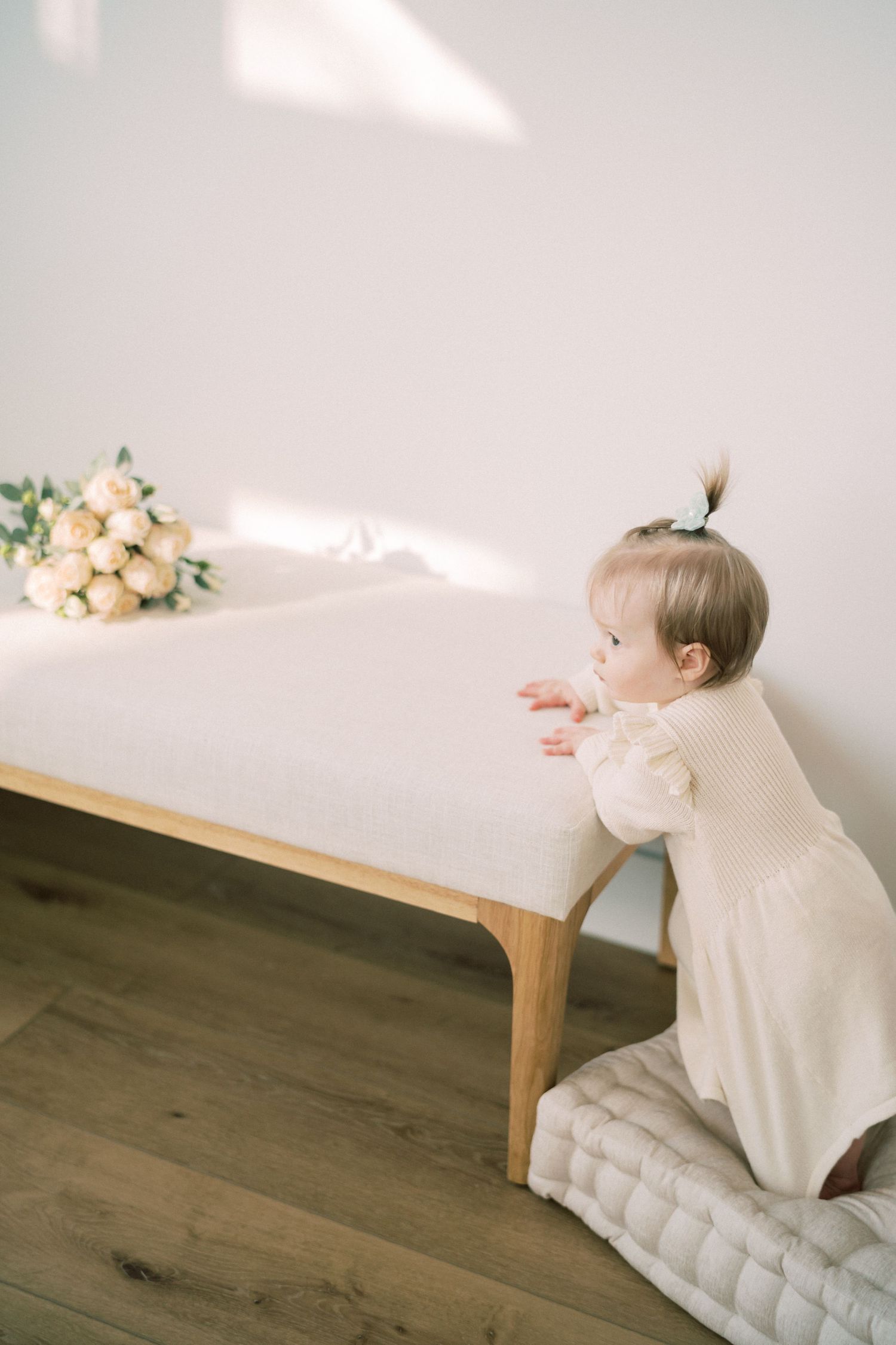 Small child in a white dress peers over the edge of a wooden table with flowers on top and cushion underneath.