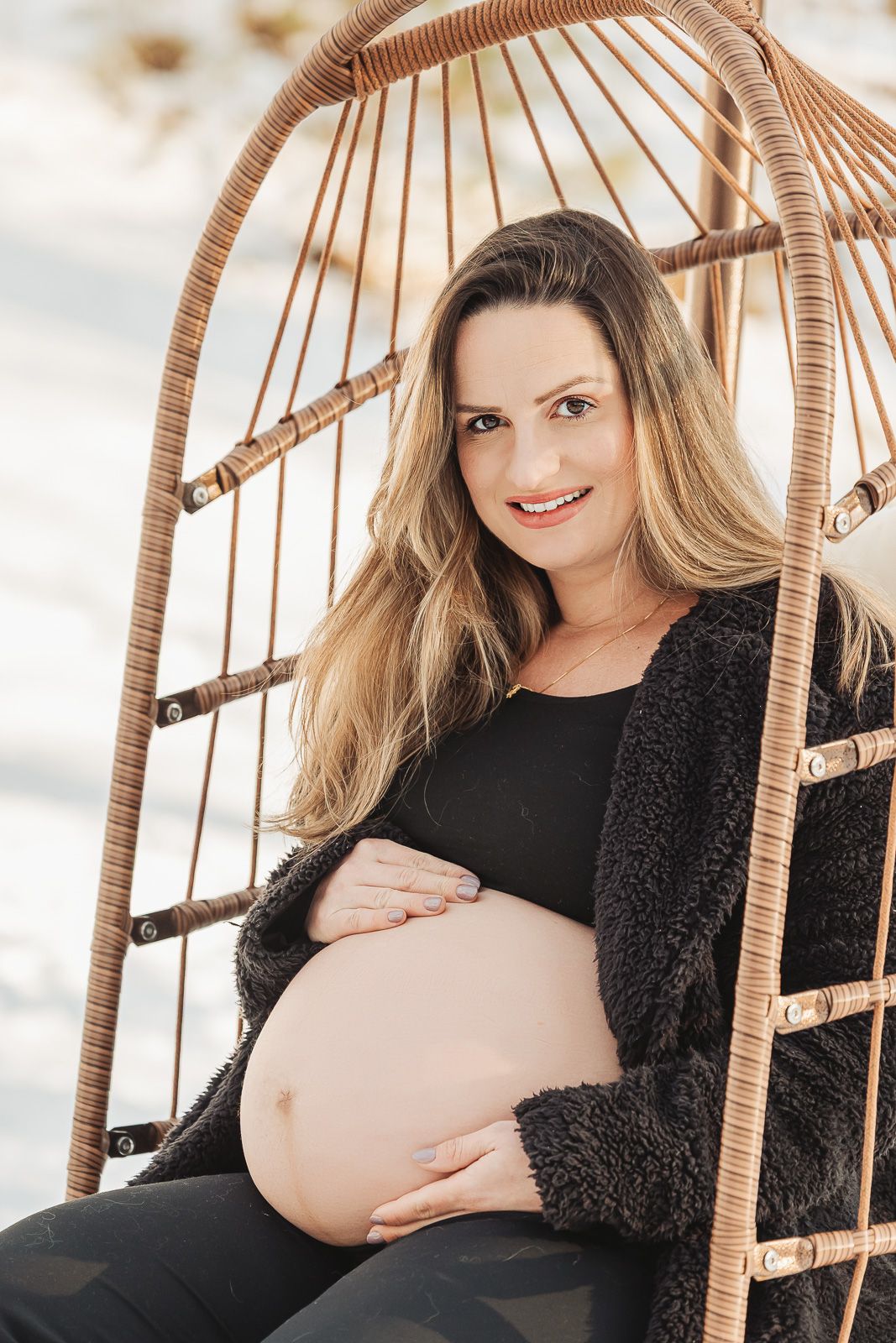 Expectant mother relaxes in wicker hanging chair wearing black outfit against snowy winter background.