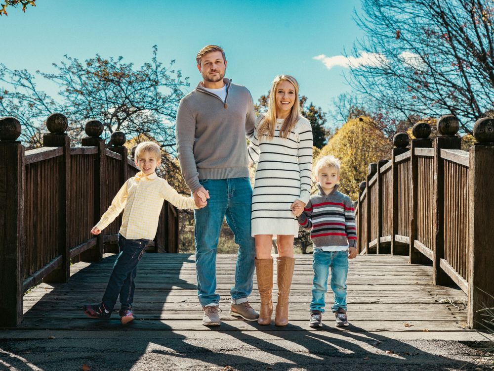 Family with two young children stands together on a wooden bridge against blue sky and fall foliage.