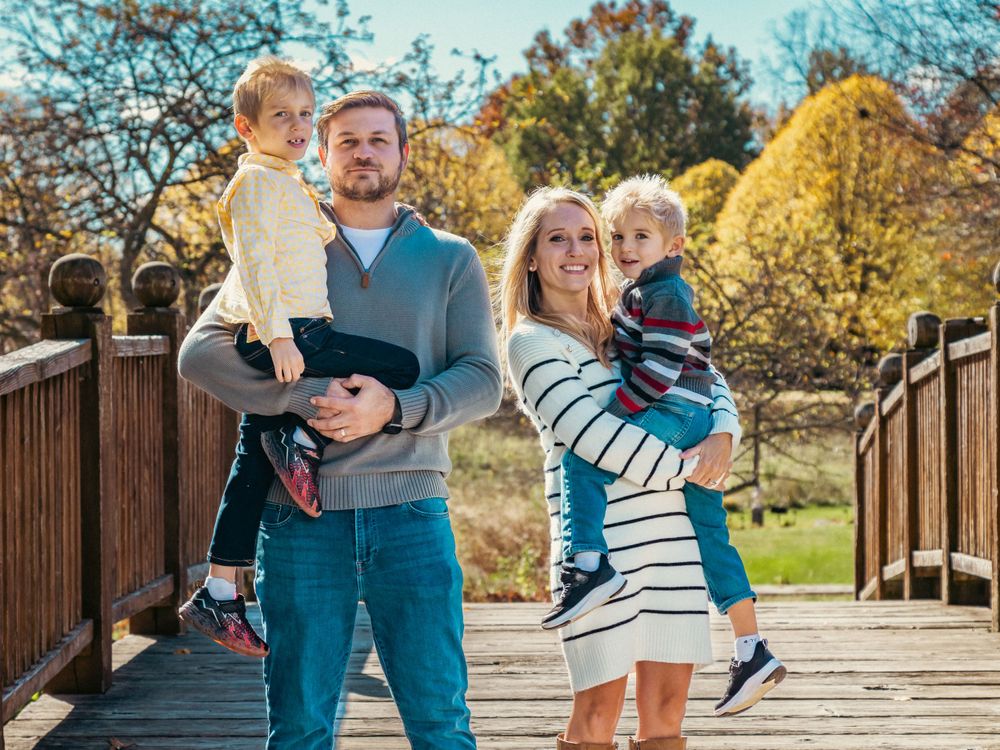 Parents hold their children while posing on wooden bridge surrounded by autumn trees.