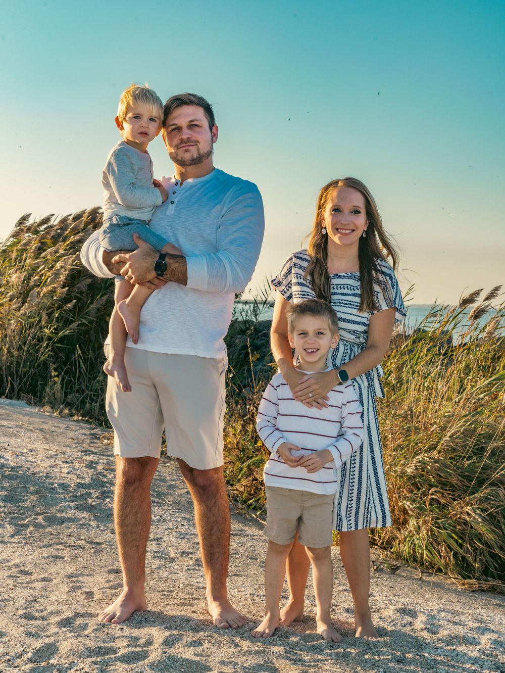 Family poses together on sandy beach path with sea oats at sunset.