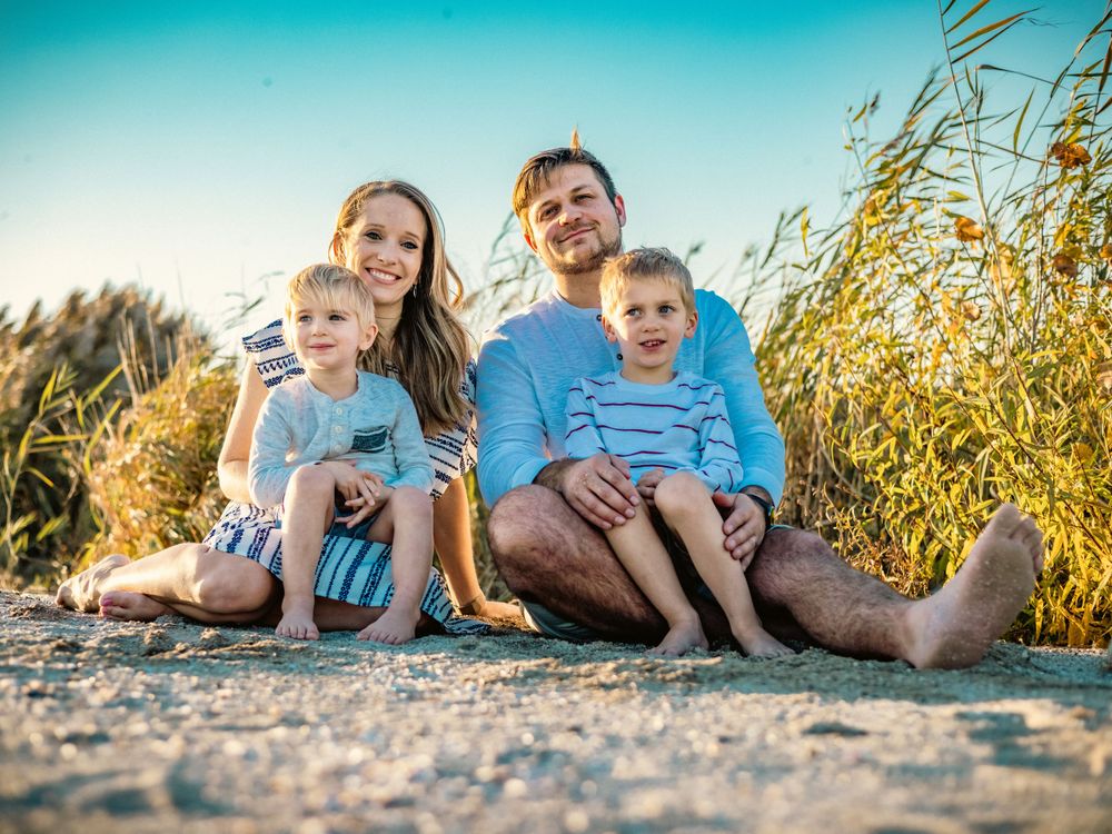 Family sits together on sandy beach with tall grass and turquoise sky background.