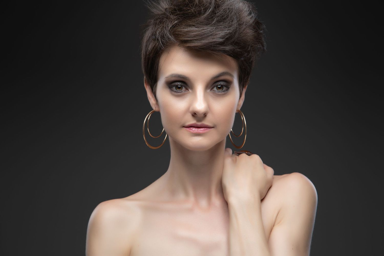 Professional portrait studio photo with dramatic lighting featuring short dark hair, silver hoop earrings and bare shoulders.
