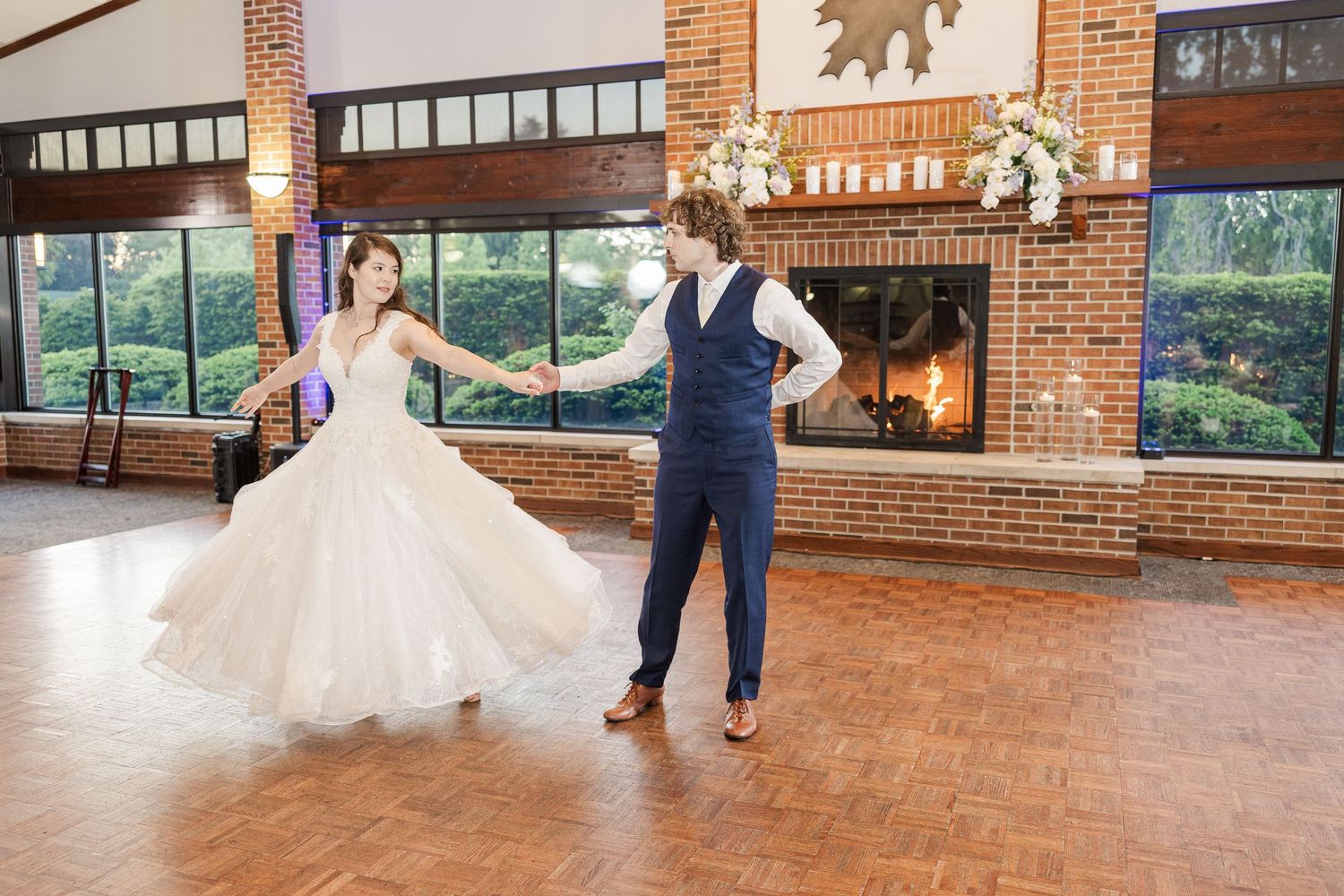 At an indoor wedding reception venue, a couple dances on a wooden floor near a rustic brick fireplace.