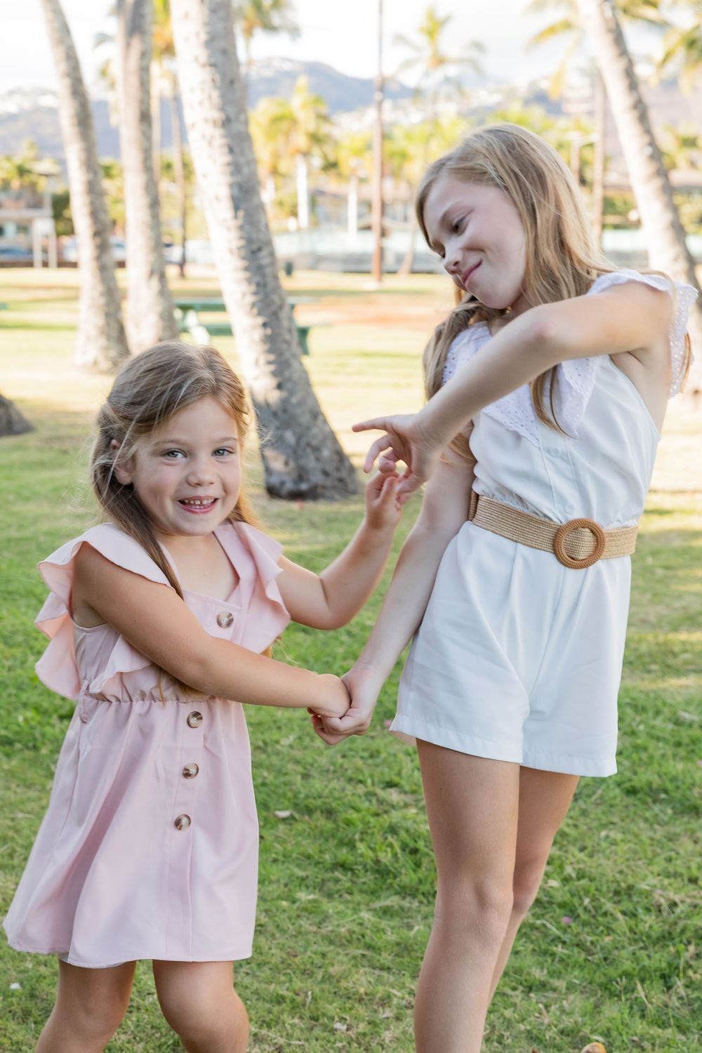 Two people in summer dresses share a playful moment together in a sunny park with palm trees in the background.