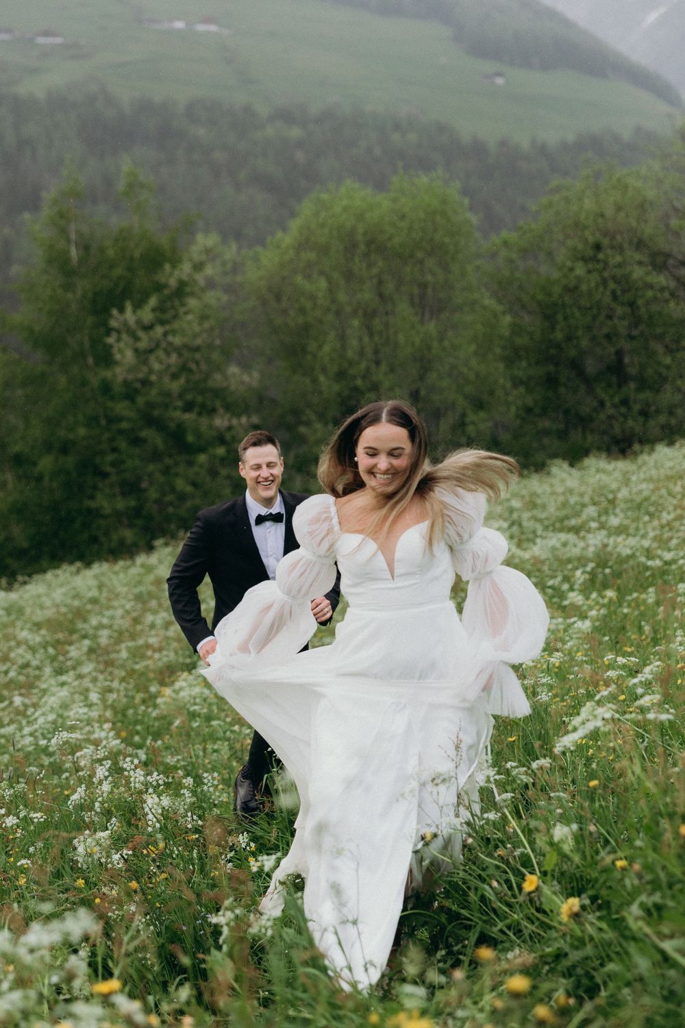 Wedding couple plays in a wildflower meadow surrounded by mountains and forest as her white gown flows in the breeze.