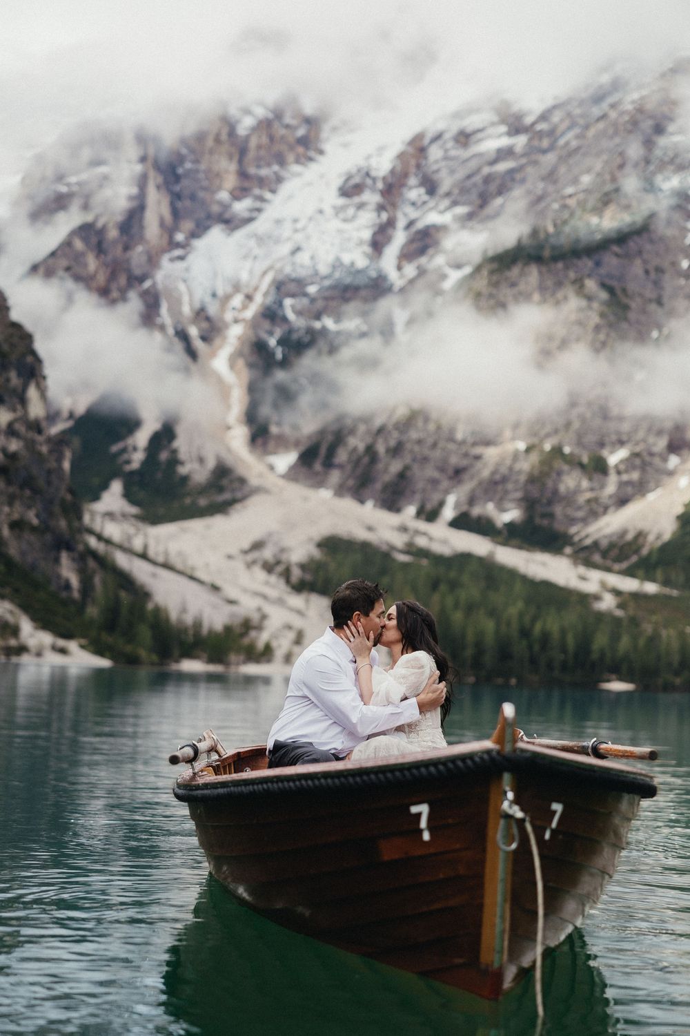 A wooden boat floats on a turquoise alpine lake with snow-capped Dolomite mountains and misty clouds in the background.