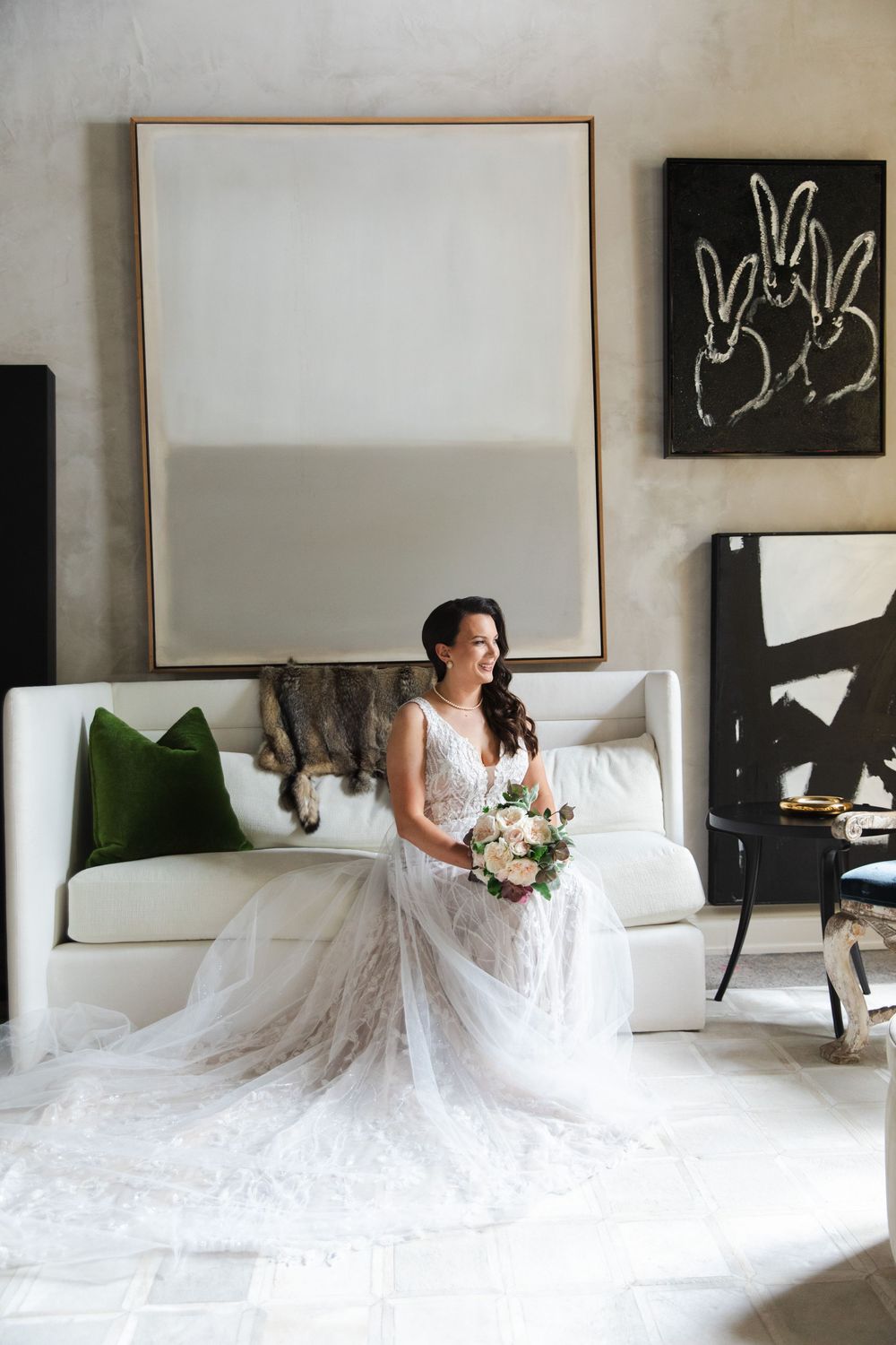 A bride in a flowing white wedding gown sits elegantly on a modern white sofa against a minimalist gallery wall.