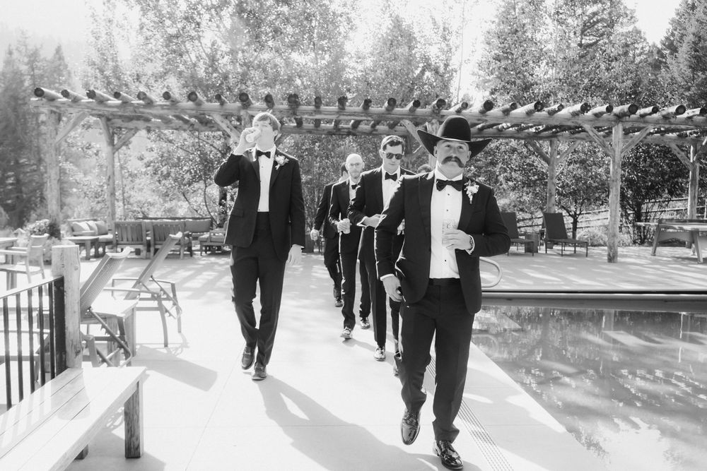 A black and white photo of groomsmen walking in line along a garden path under a pergola structure.
