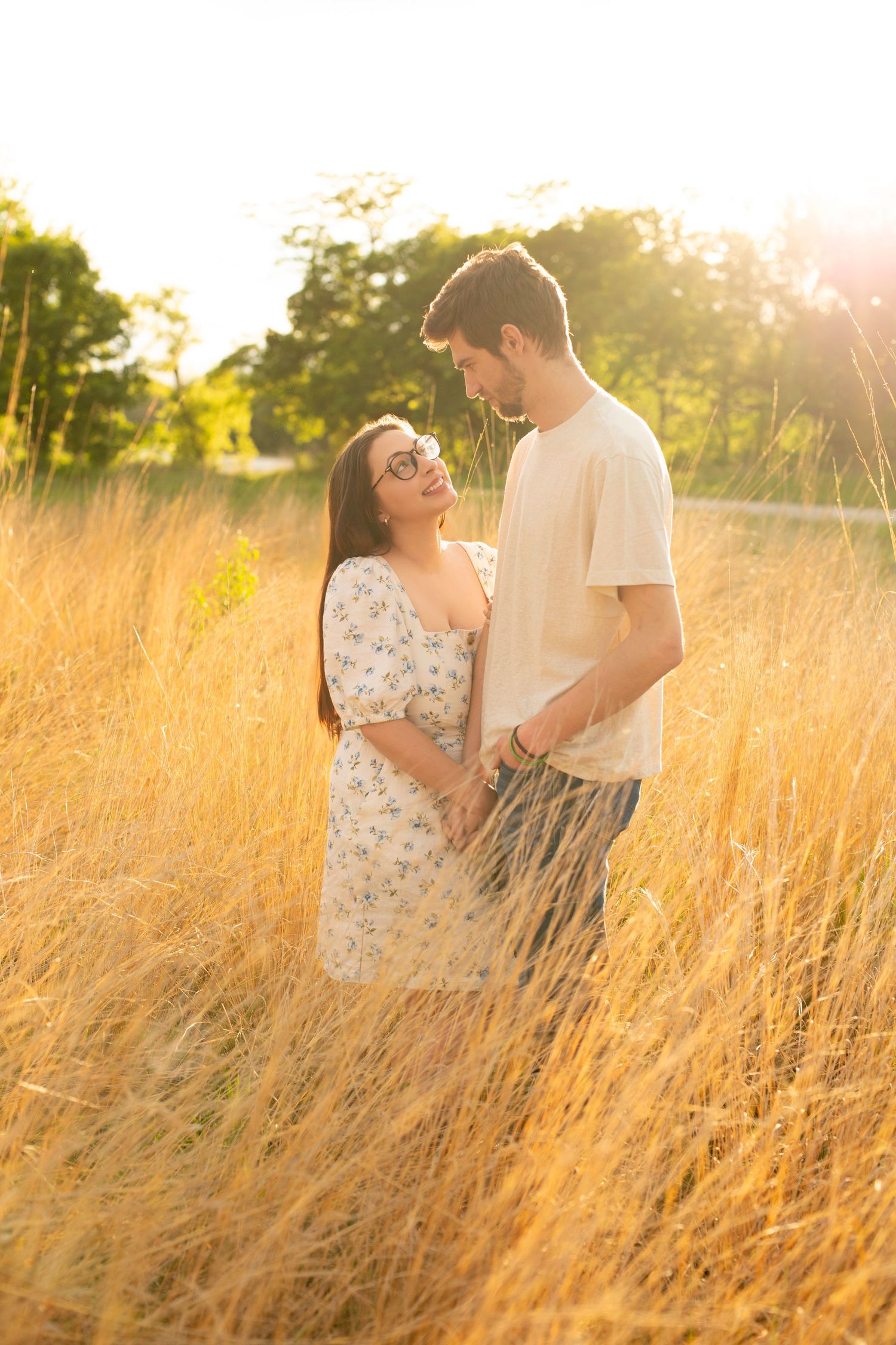 A couple stands embracing in a golden field of tall grass during sunset, creating a warm romantic atmosphere.