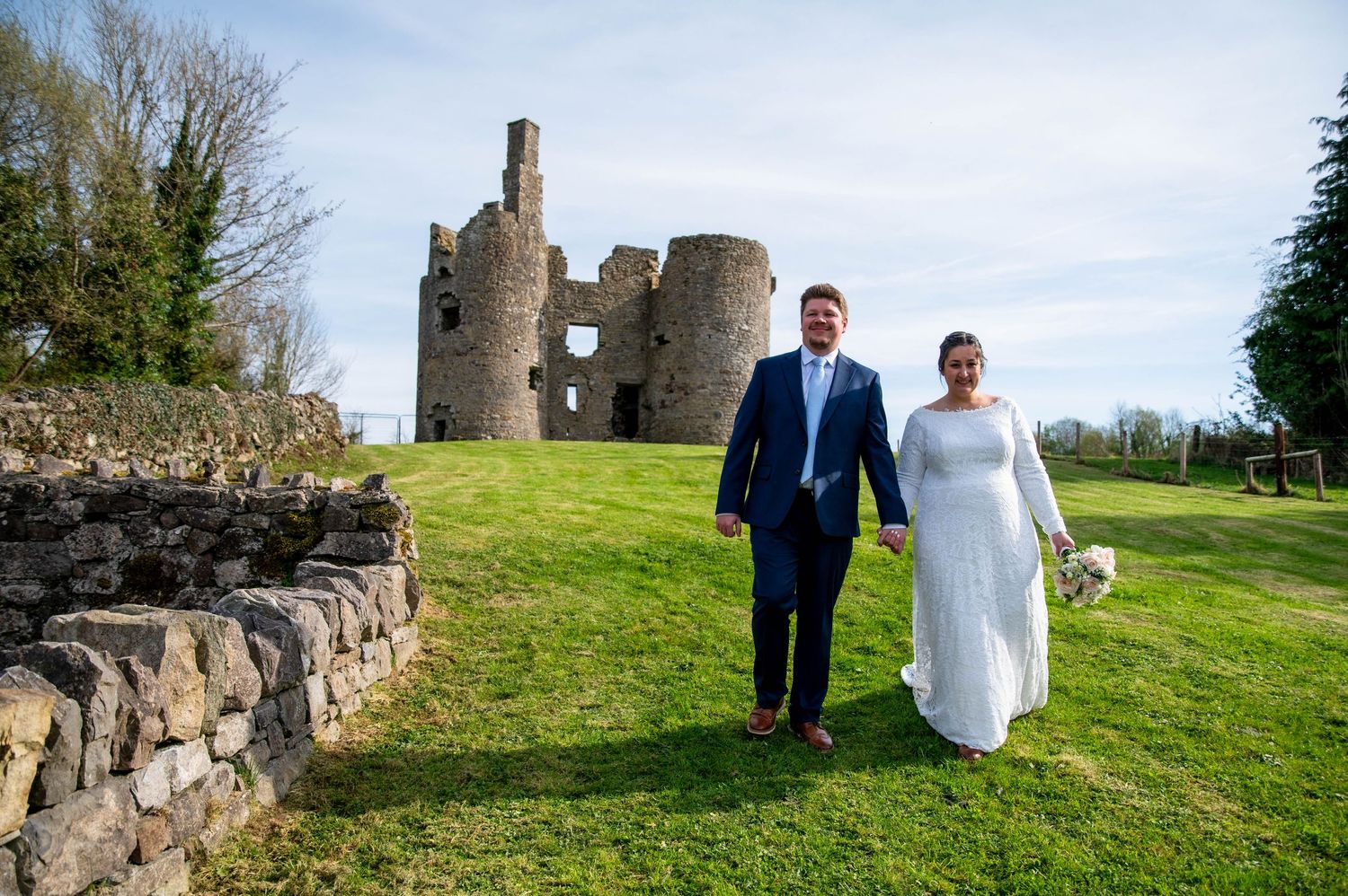 A couple stands on a grassy hill in front of the ruins of a stone castle tower under blue skies.