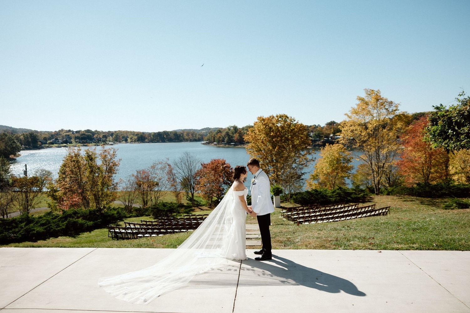 A couple in wedding attire stands on a paved area overlooking a scenic lake and autumn foliage.