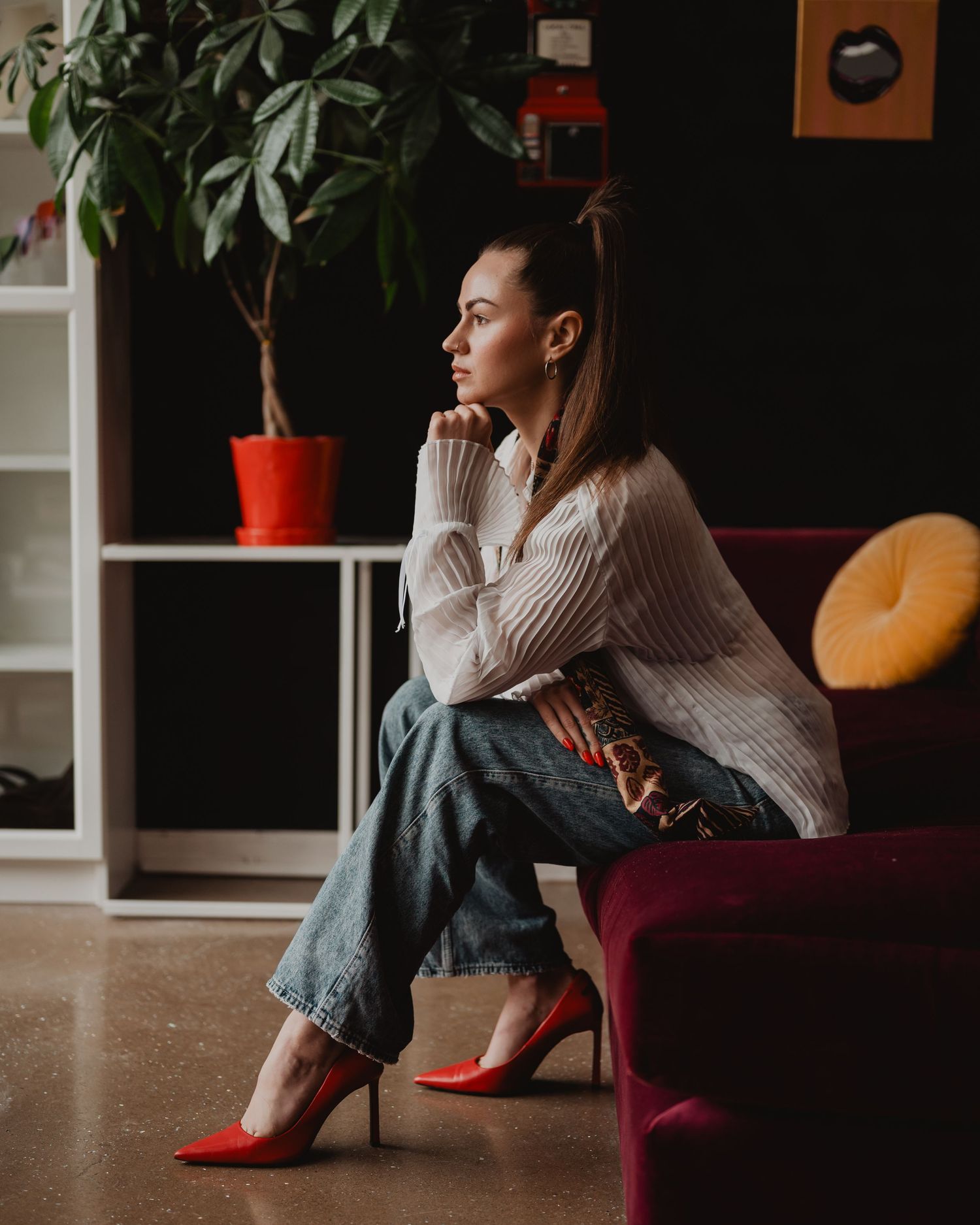 Person in casual sweater and jeans sits contemplatively on maroon chair near houseplants in dimly lit interior space.