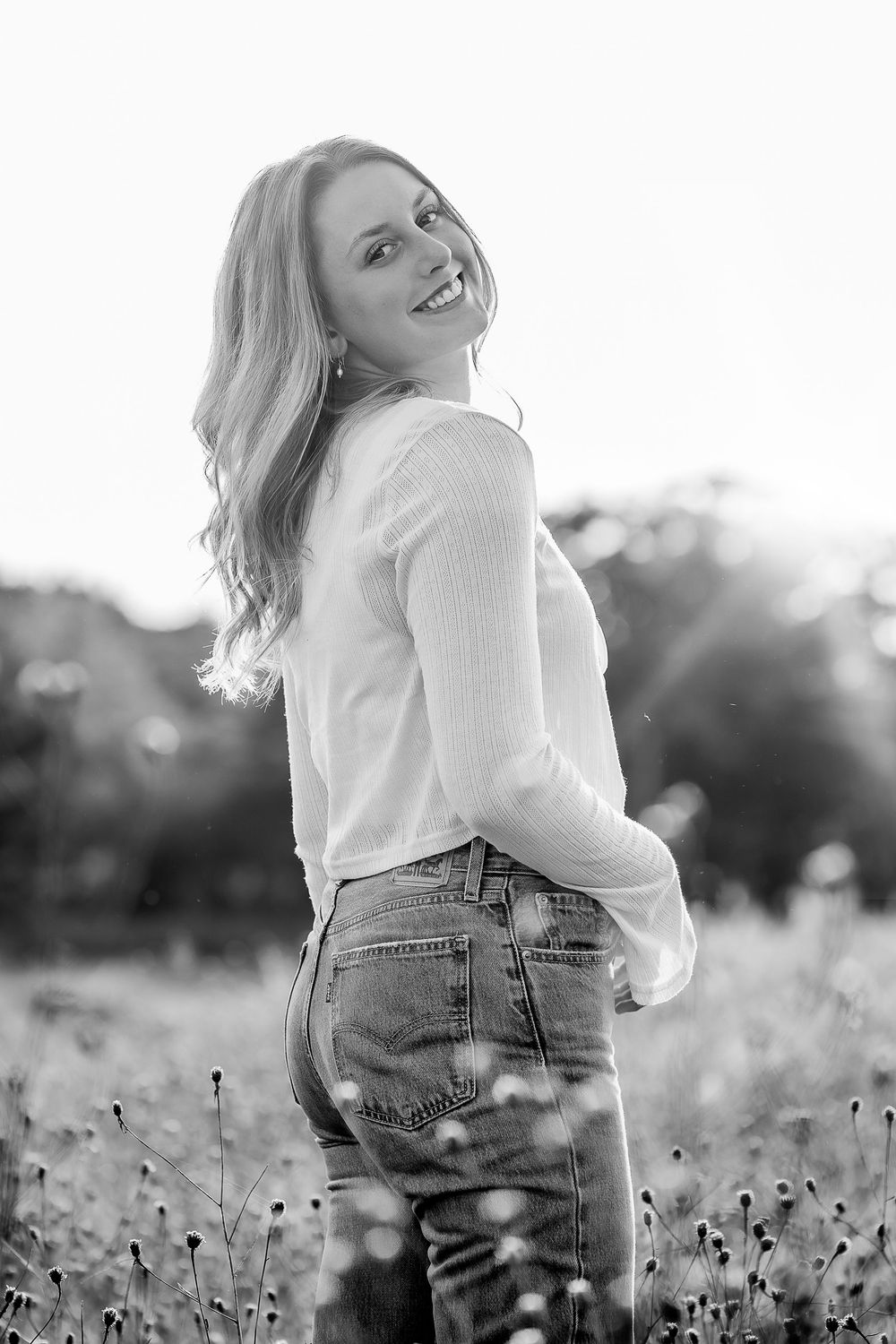 Black and white portrait of a person in a white sweater and jeans standing in a wildflower field at sunset.
