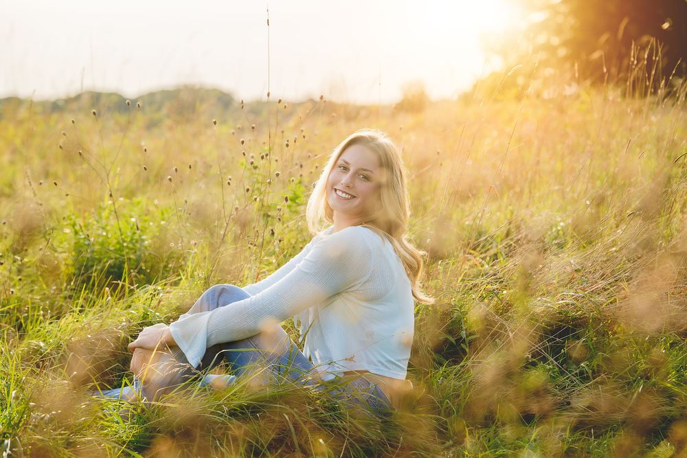 Someone in a white sweater sits in a sunlit meadow with tall grass and wildflowers during golden hour.