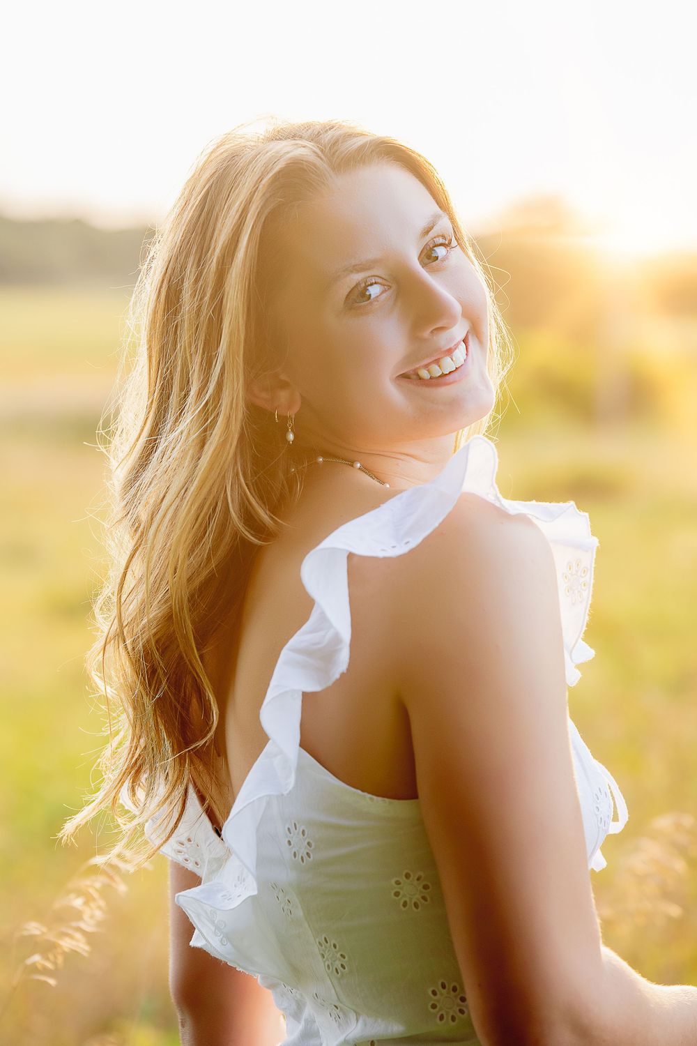A person in a white ruffled dress smiles warmly in a sunlit field during golden hour.