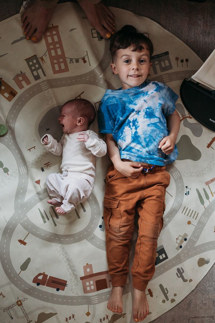 Two children lying on a round play mat featuring a city road design with buildings and vehicles in muted colors.