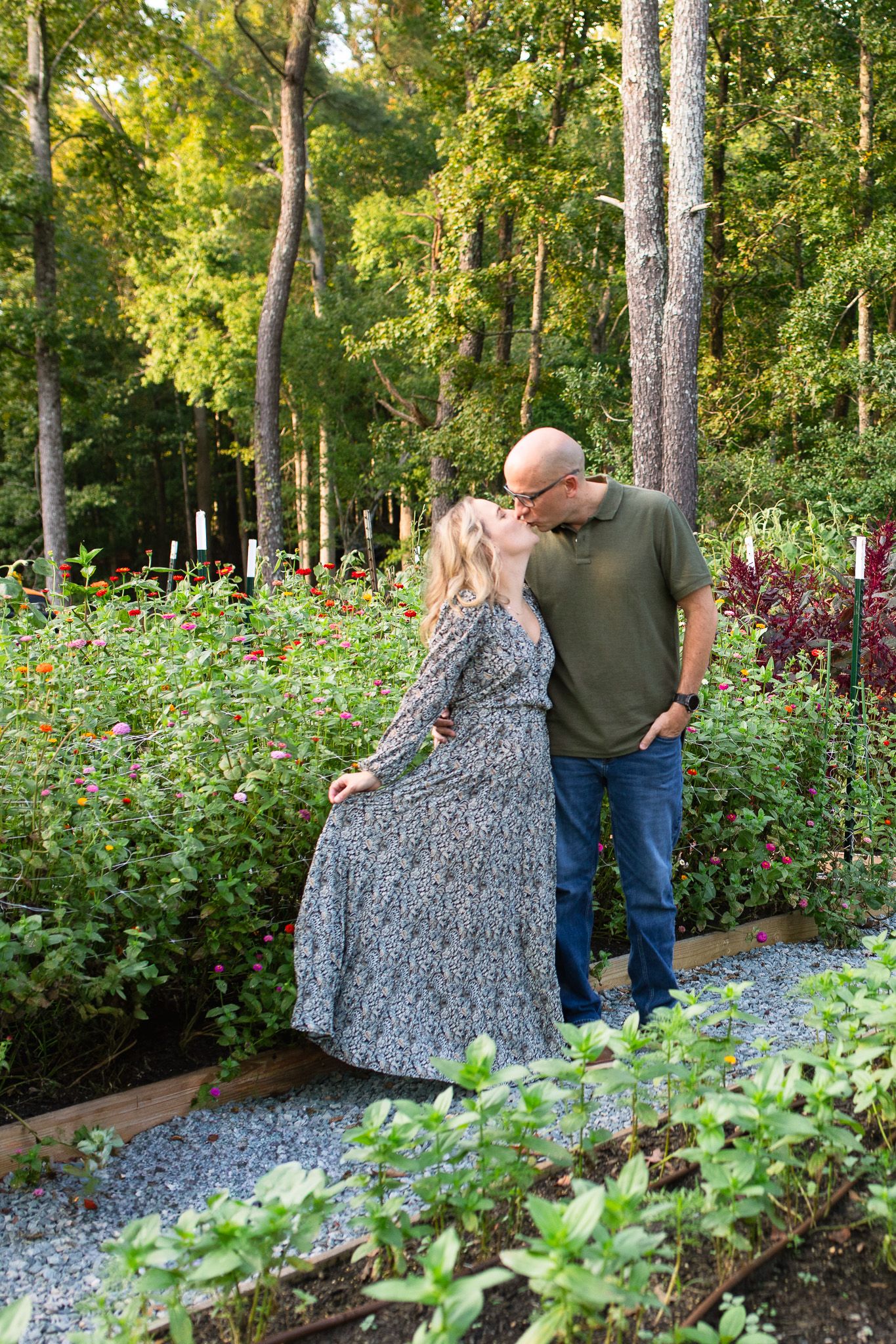 A couple shares a tender moment on a gravel path surrounded by lush garden flowers and tall trees at Polly's Petal Patch