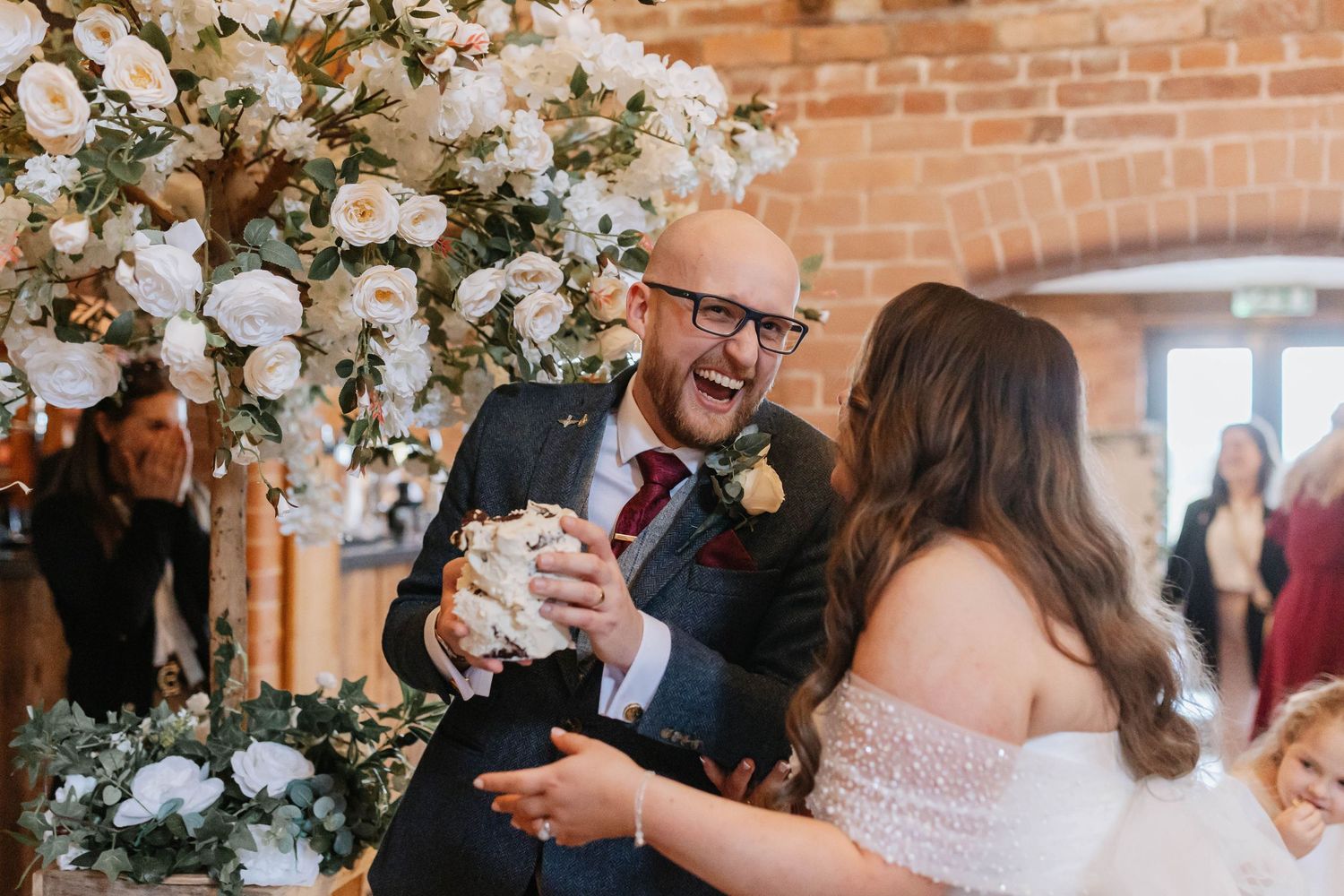 Guests sharing joyful reactions during wedding reception at brick venue adorned with white floral arrangements.