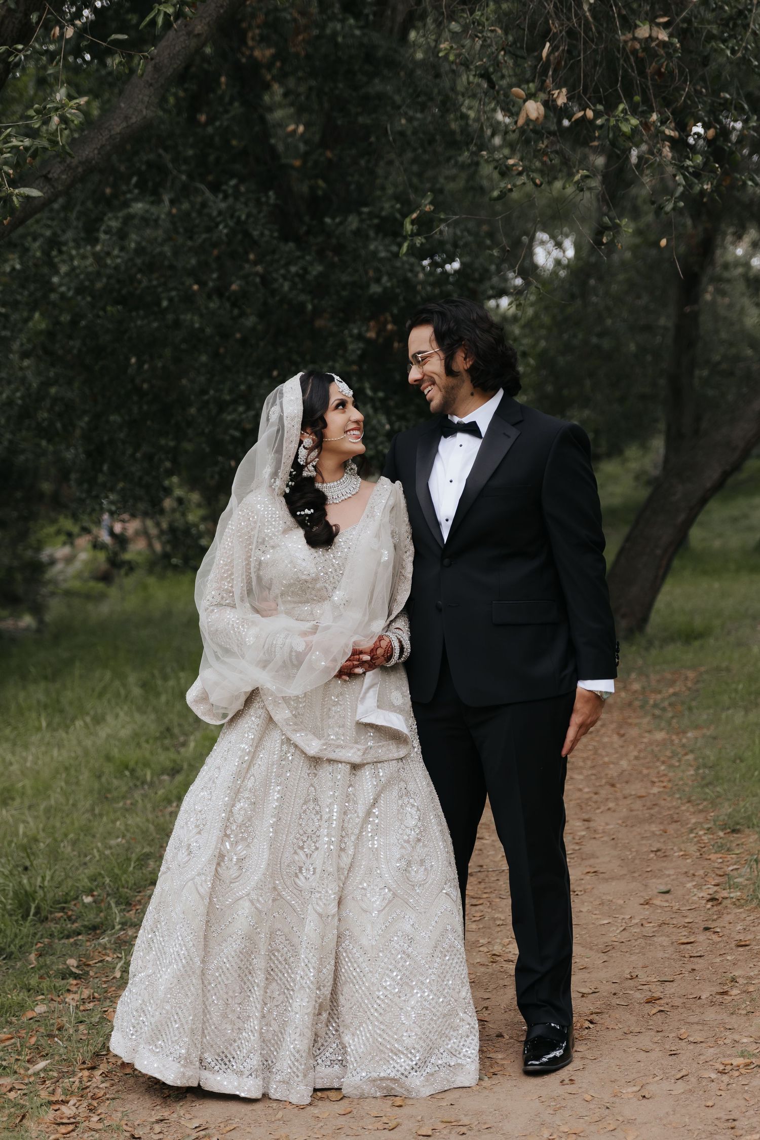 Newlyweds in traditional wedding attire walk along a woodland path while gazing at each other.