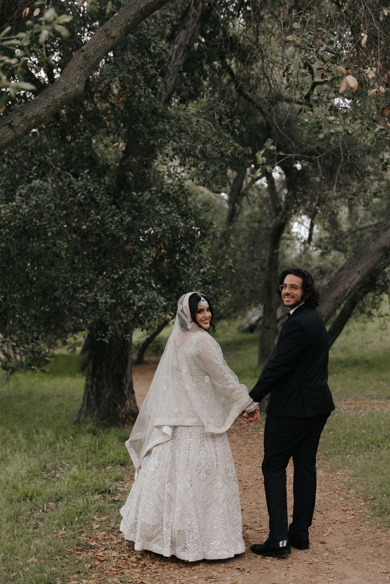 Wedding couple holds hands on a natural path with autumn trees in the background.