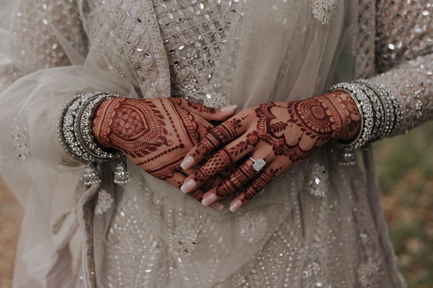 Hands adorned with traditional henna designs rest gently on a white lace wedding dress in soft, warm lighting.
