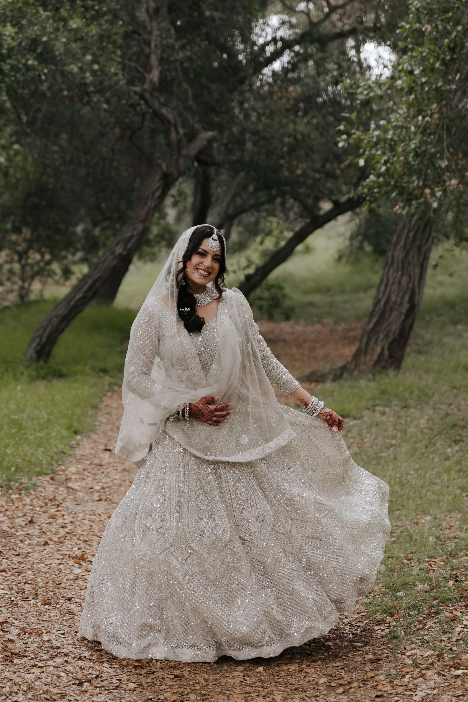 A bride in an ornate white lace wedding dress twirls on a tree-lined path, showcasing the flowing layers of her gown.