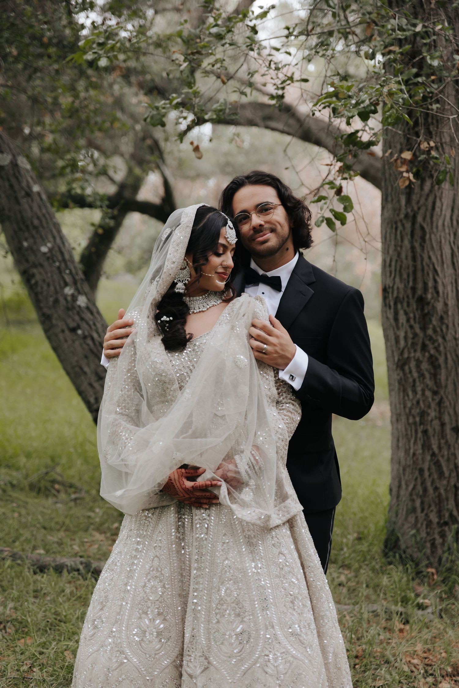 Romantic wedding portrait of couple embracing under trees in formal attire.