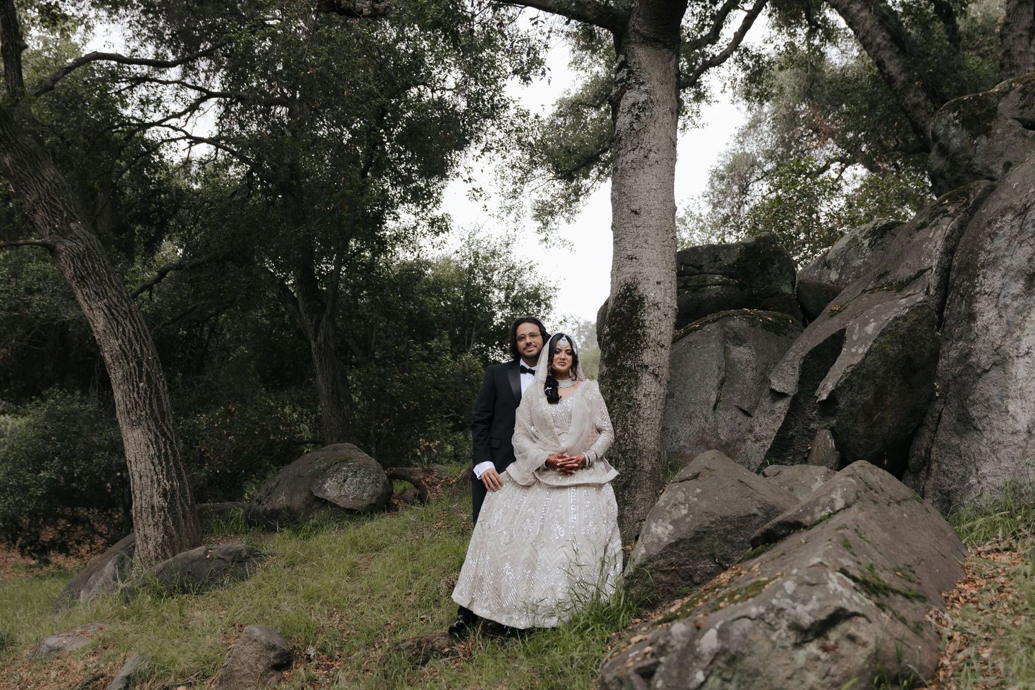 Wedding couple poses on large rocks surrounded by trees in a natural setting.