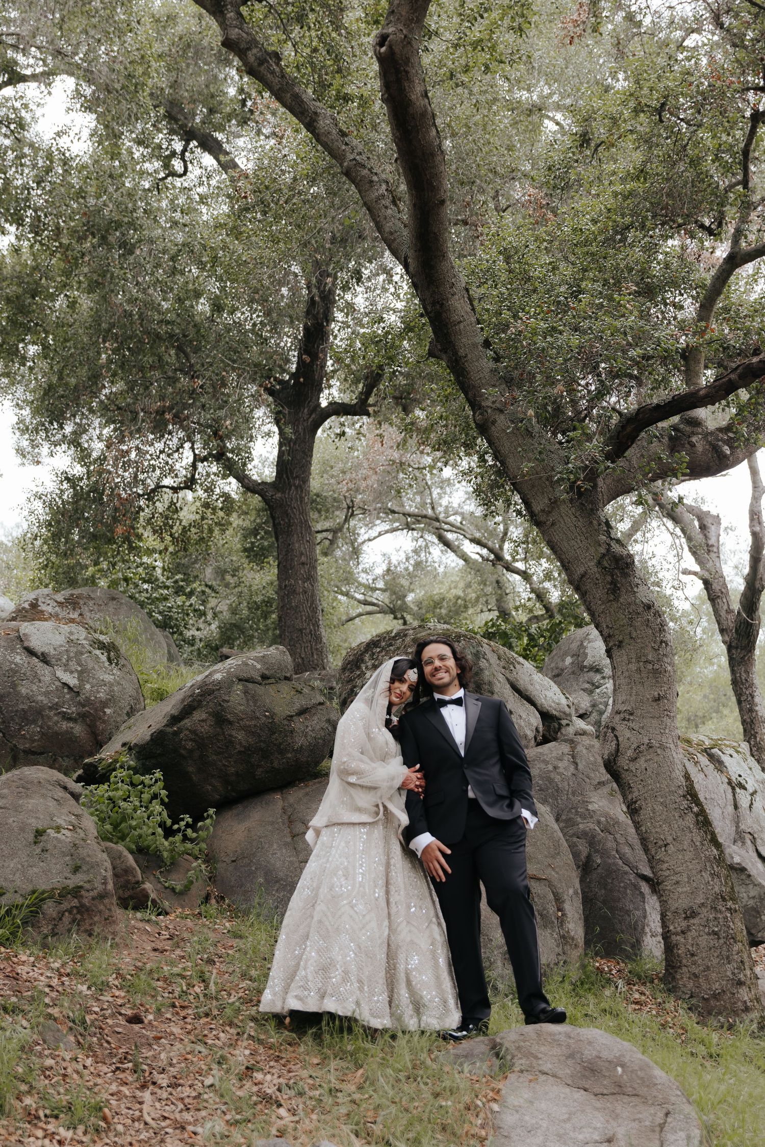 A couple in wedding attire poses romantically among large boulders and trees in a rustic outdoor setting.