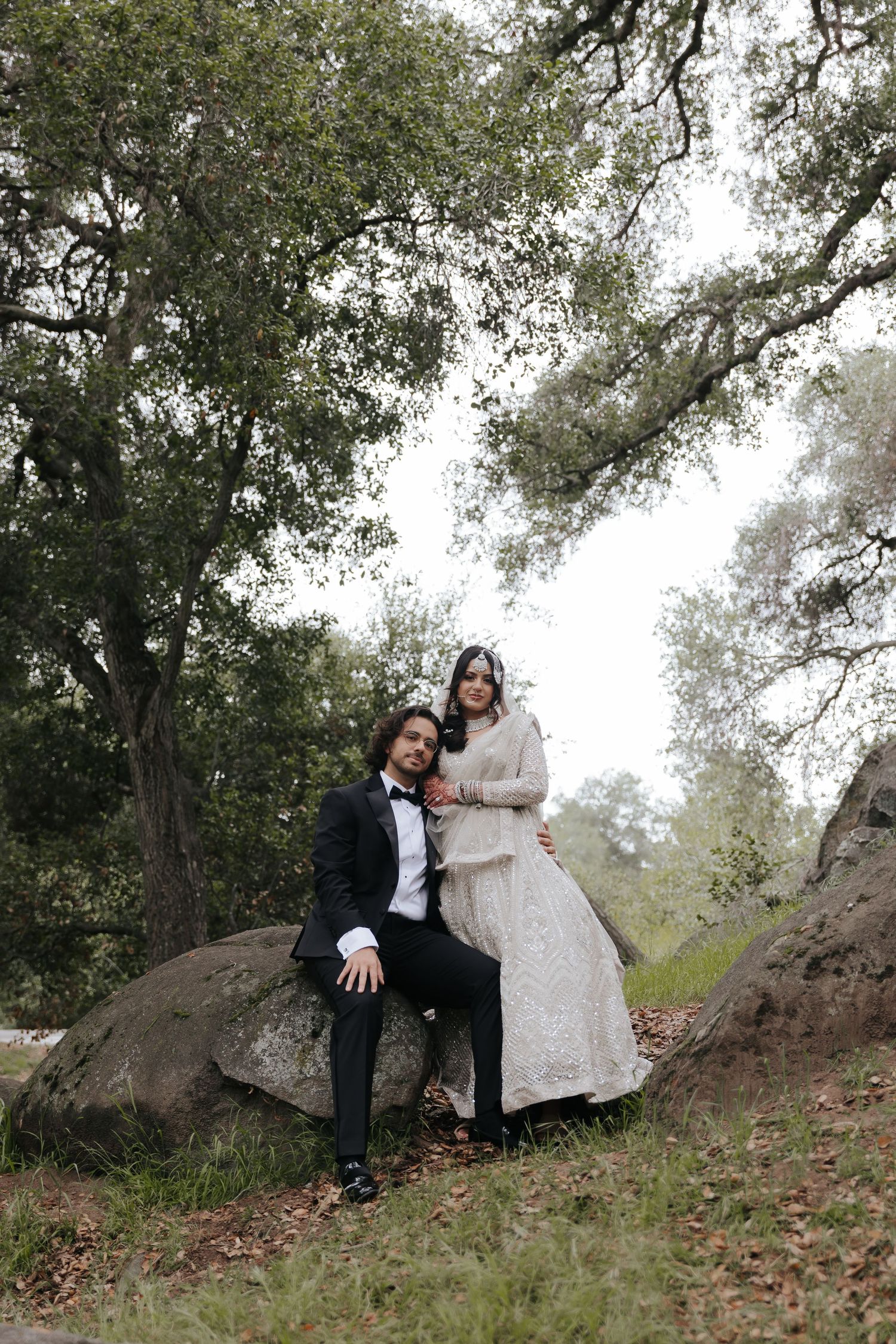 A couple poses together in a romantic outdoor setting with trees and rocks in a natural woodland environment.