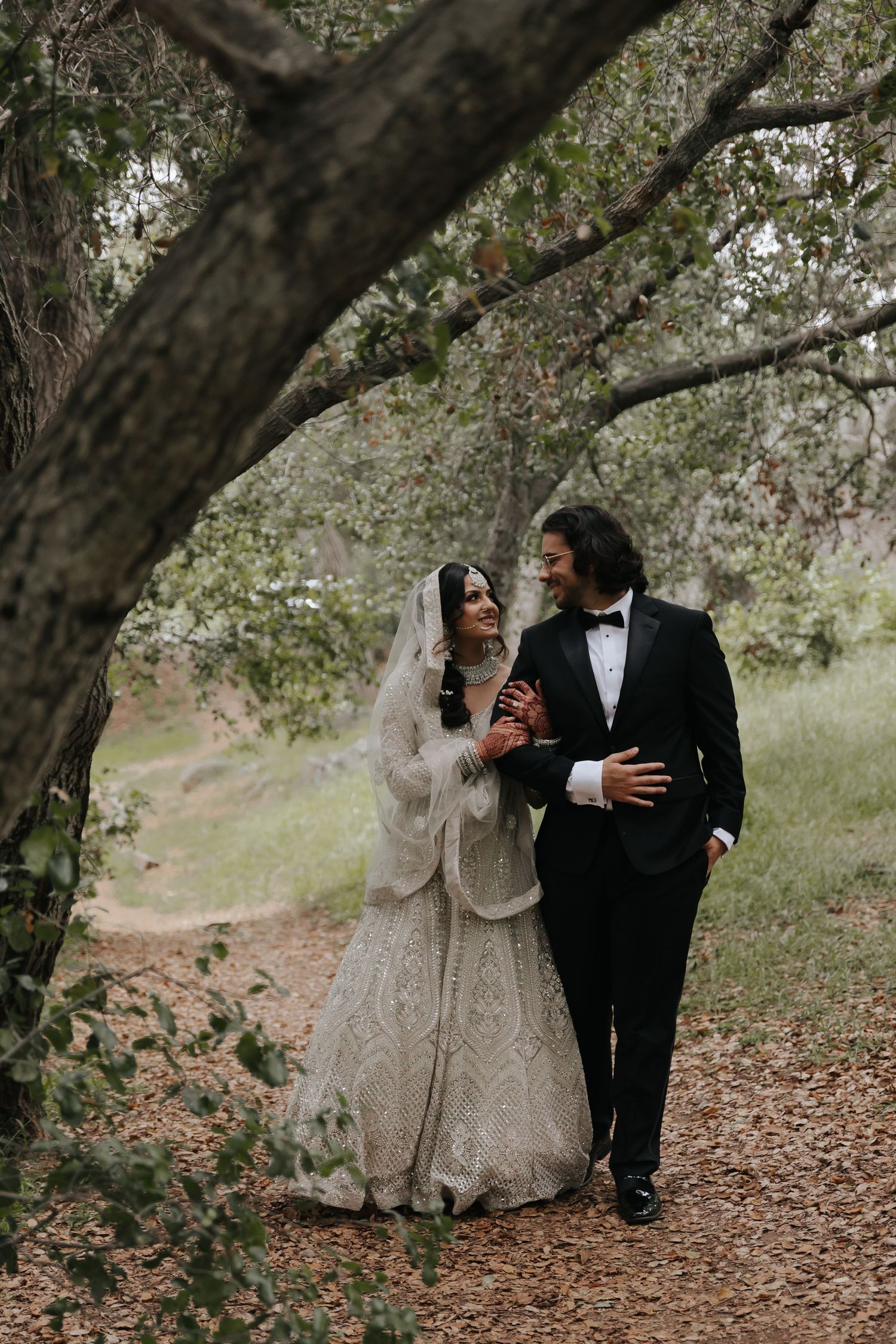 A romantic wedding couple walks together on a tree-lined path with falling flower petals on a misty day.
