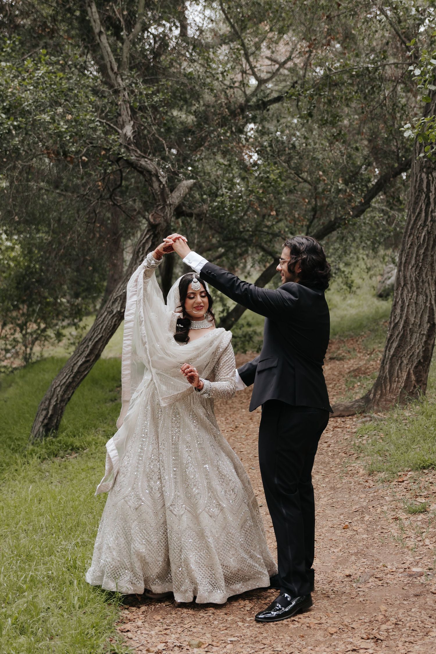 A groom helps adjust his bride's veil during their outdoor wedding photoshoot among olive trees.