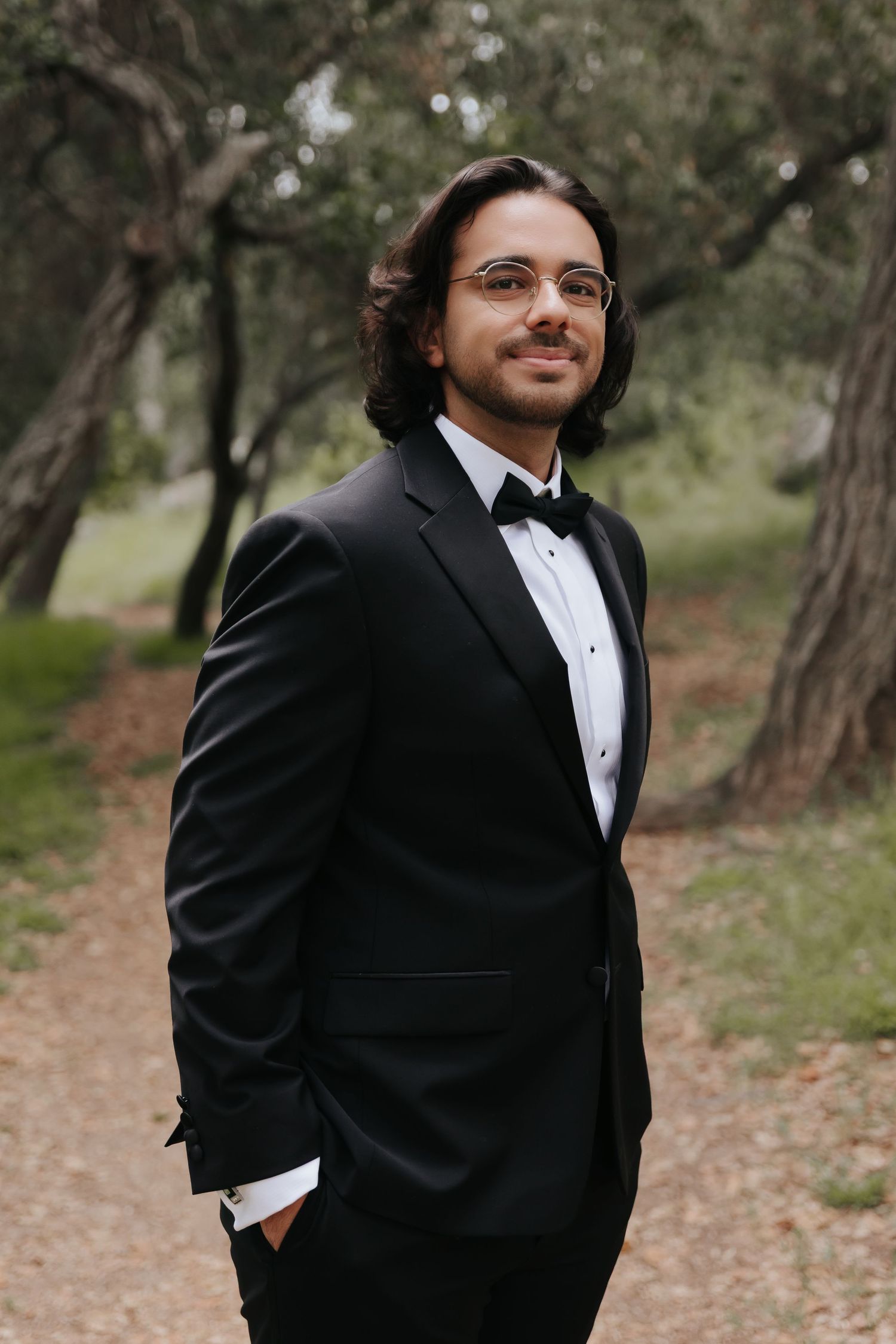A groom in a classic black tuxedo poses on a wooded path with natural scenery.