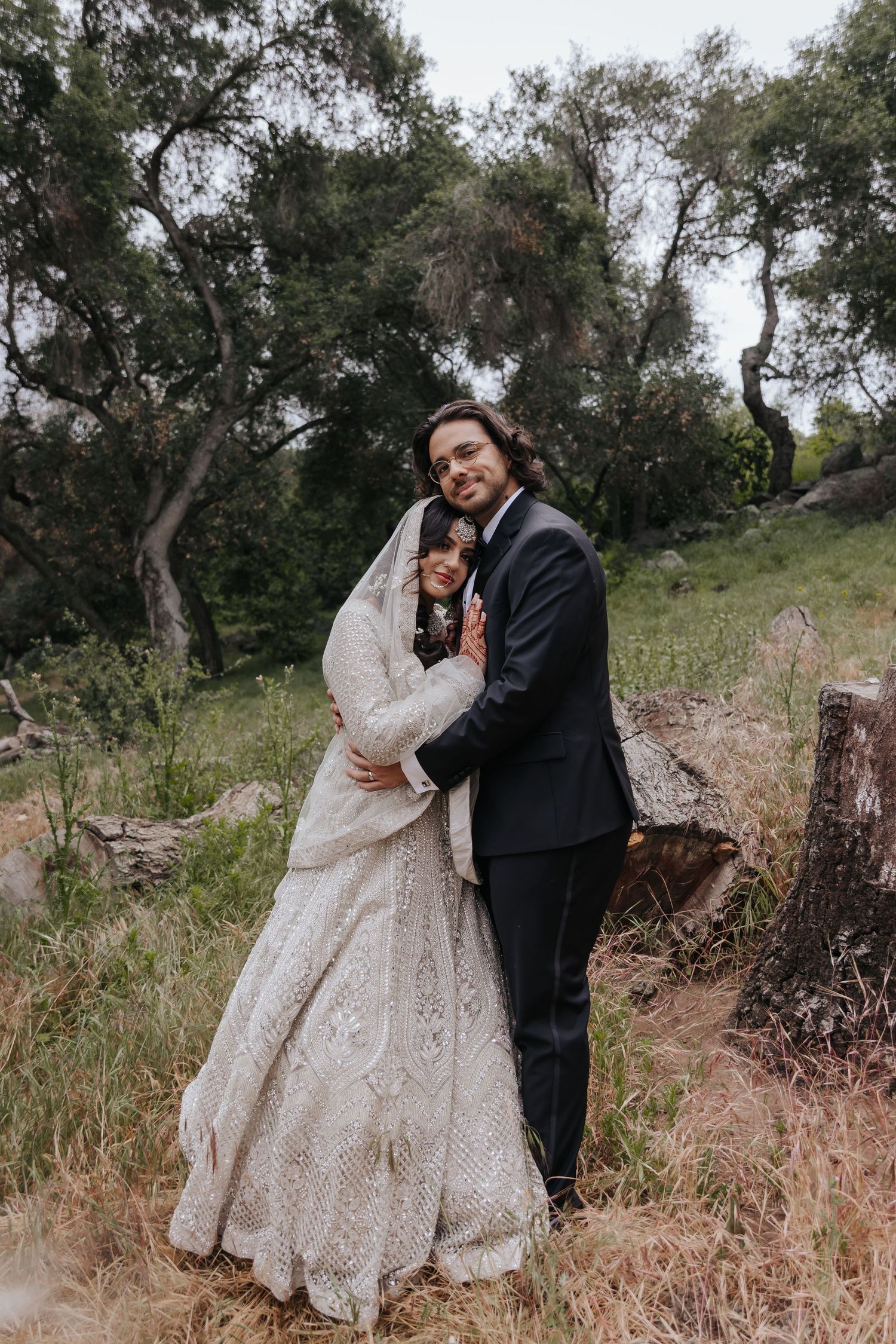 A newlywed couple embraces on a rustic outdoor path surrounded by trees.