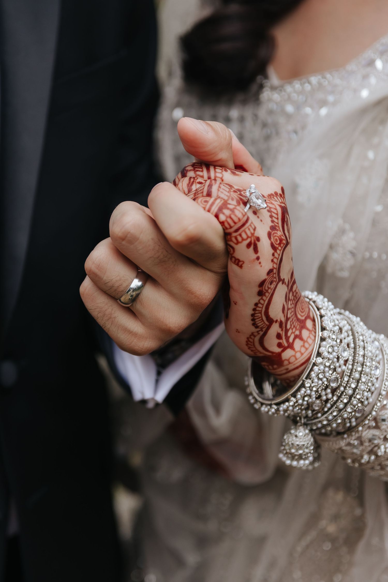 Close-up of joined hands showing mehndi designs, wedding ring, and silver bangles.