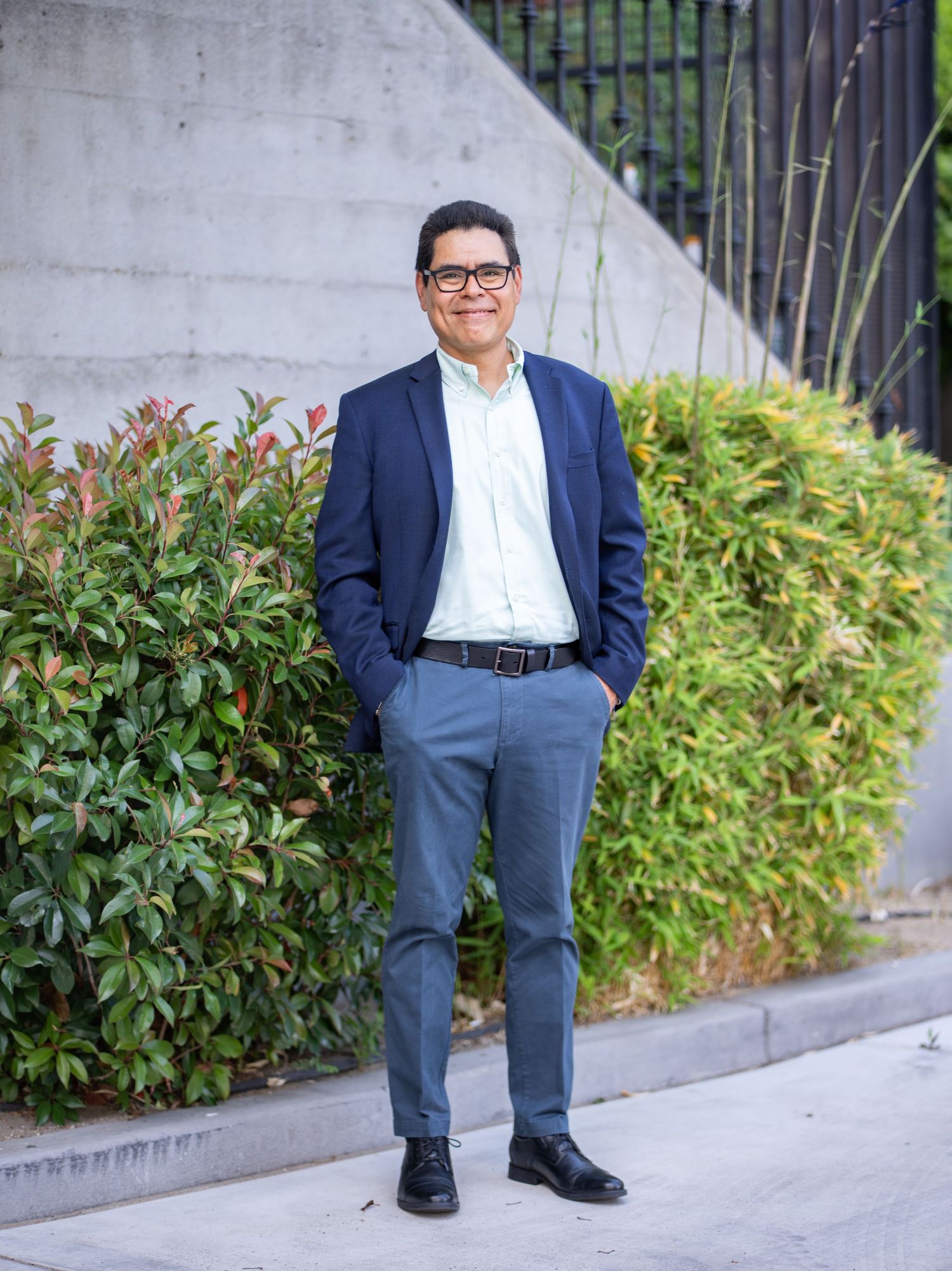 Professional portrait of someone in a navy blazer and glasses standing outdoors with greenery and architecture in background.