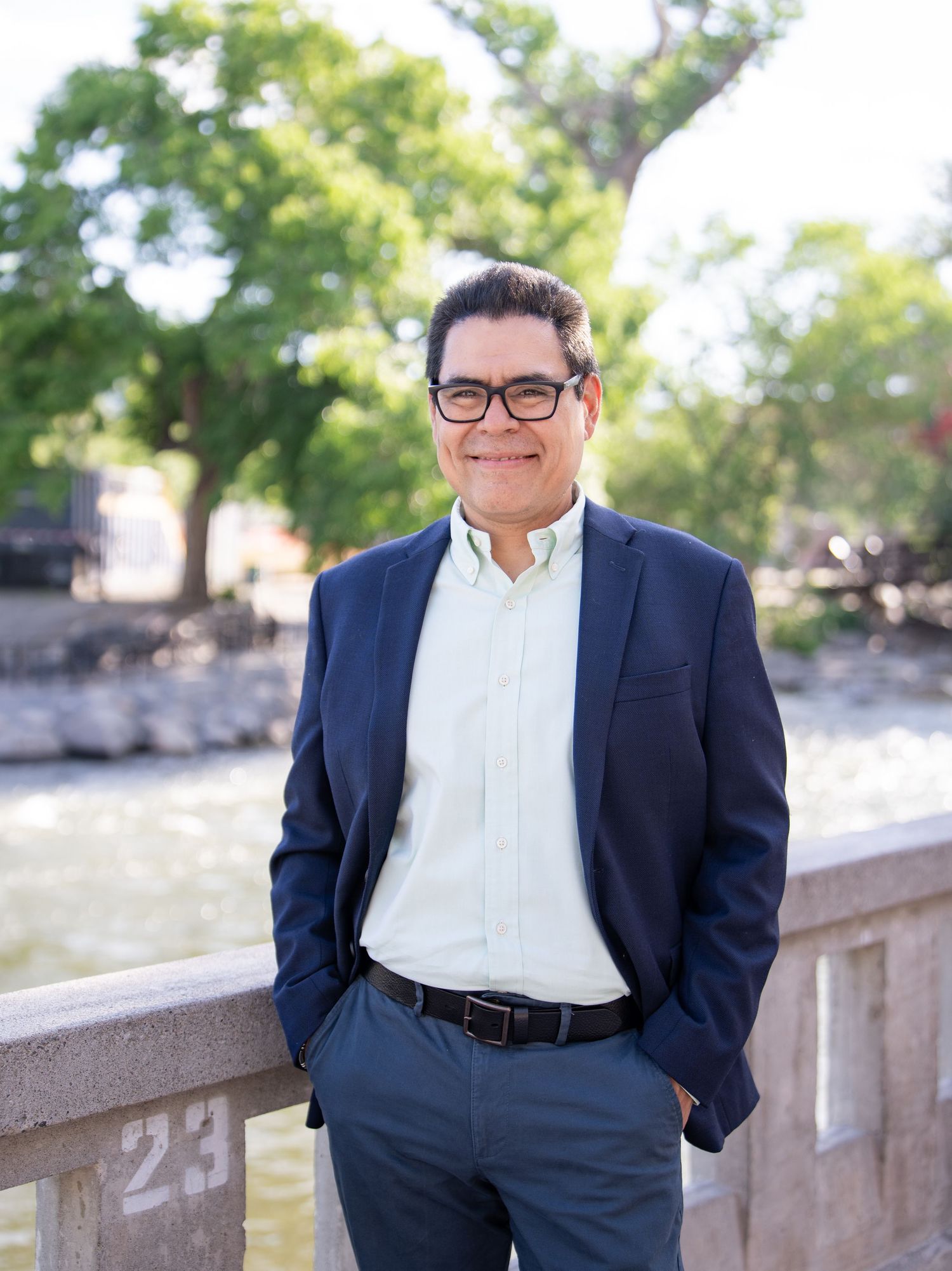 Professional wearing navy suit and glasses poses outdoors against blurred natural background and stone railing.