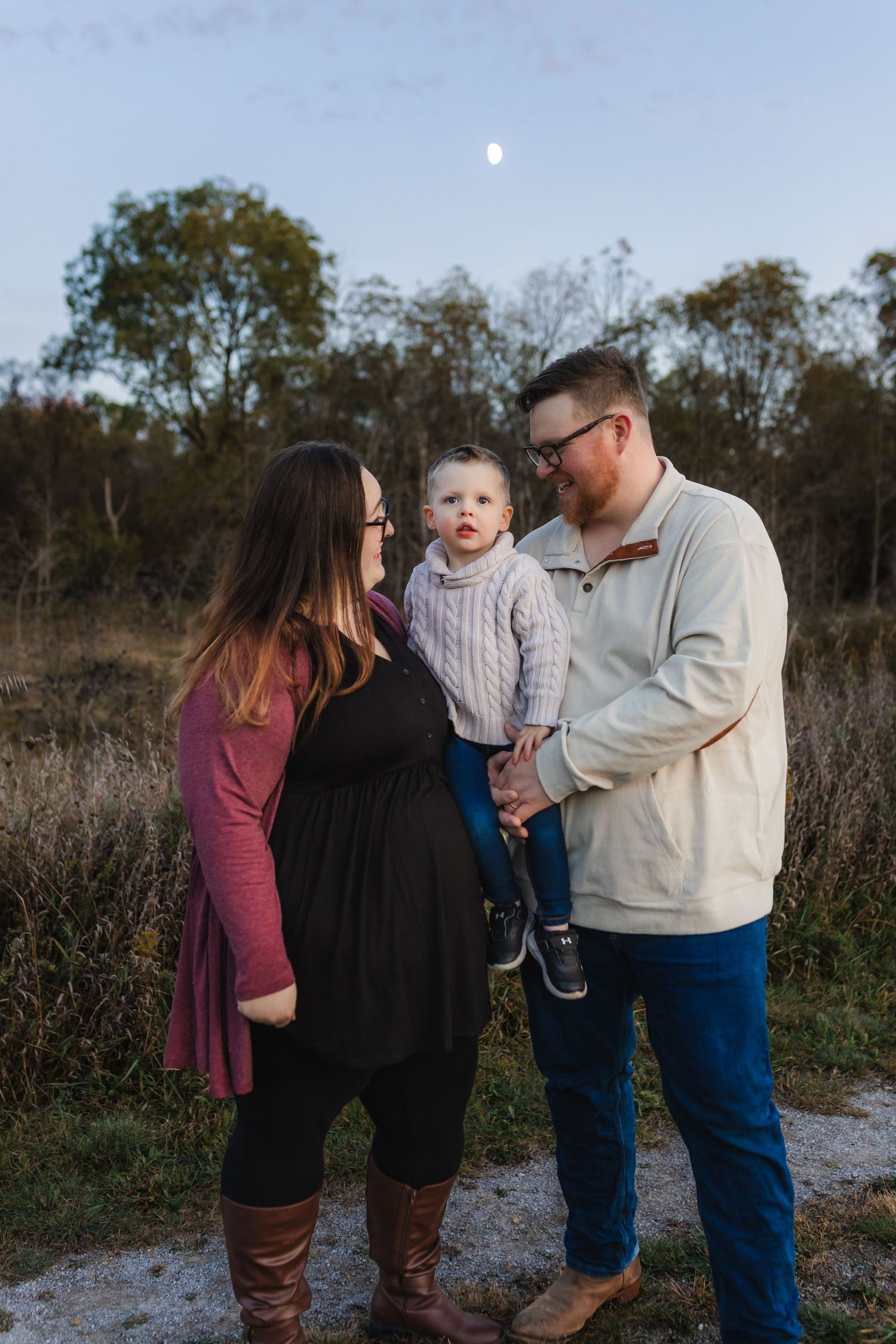 A family stands together outdoors at dusk with a moon visible in the sky and autumn trees in the background.