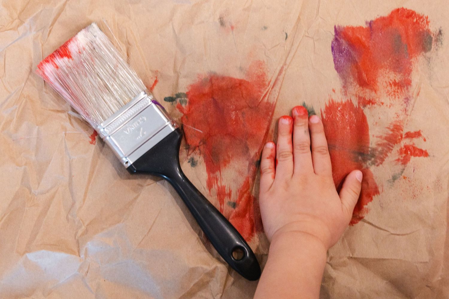 A small child's hand next to a paintbrush with red paint and strokes on brown craft paper.