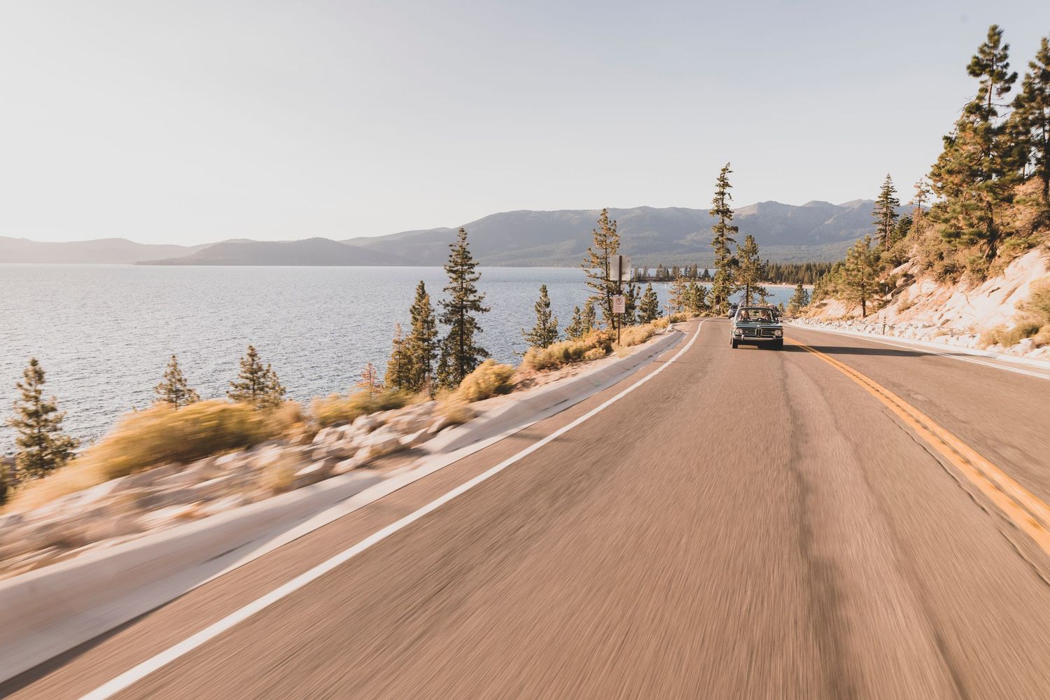 Scenic mountain road curves along Lake Tahoe with evergreen trees and snow-capped peaks in the distance.