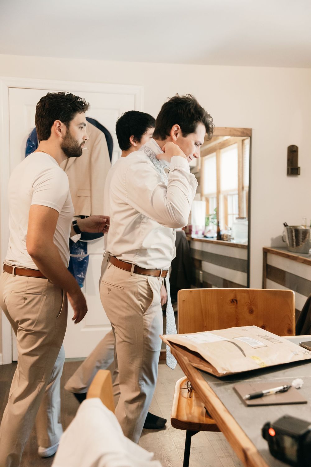 Groomsmen in light colored attire getting ready in a bright room before a wedding ceremony.