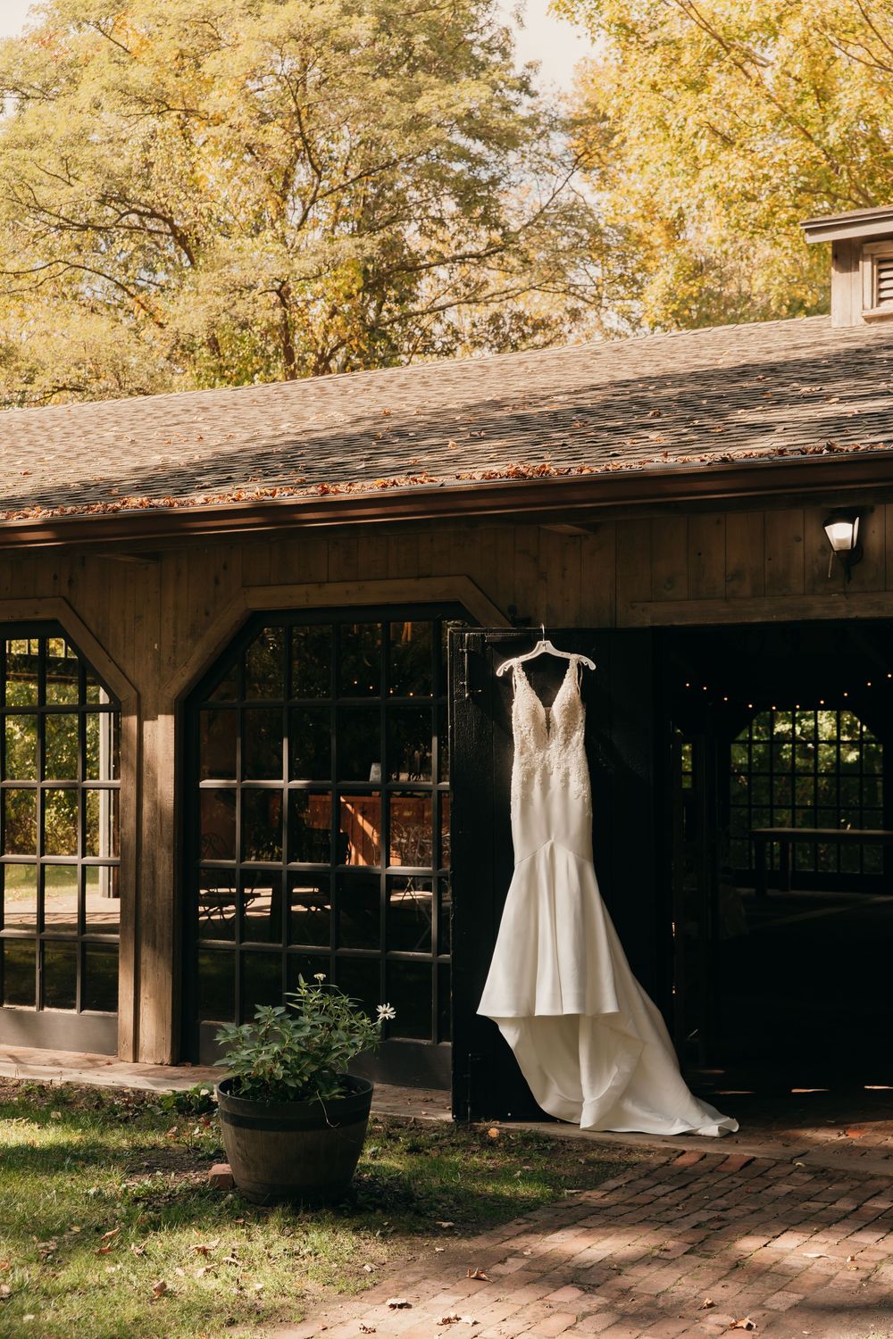 Elegant white wedding dress hanging outside a rustic wooden building with glass windows in autumn setting.