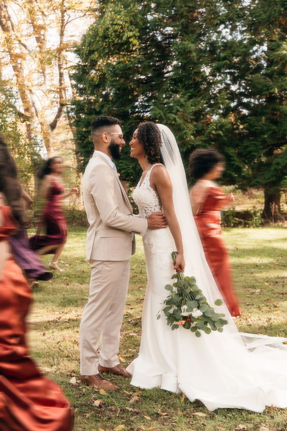 A couple embraces outdoors in elegant wedding attire with bridesmaids in rust-colored dresses.
