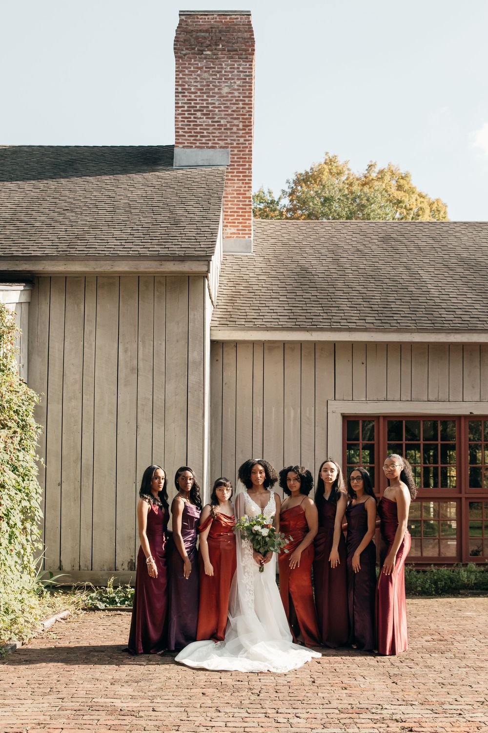 A bride in a white gown stands with her bridesmaids wearing burgundy and red dresses outside a rustic building.